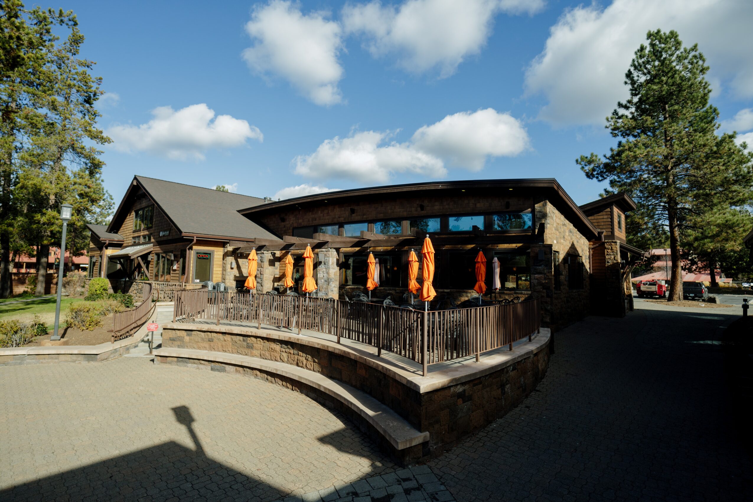 Building with curved roof and outdoor seating area, surrounded by trees and a railing, under a partly cloudy sky.