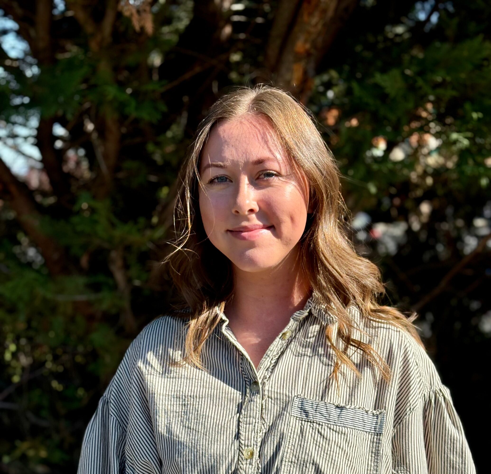 Young woman with wavy hair outdoors, wearing a striped shirt, smiling, with trees in the background.