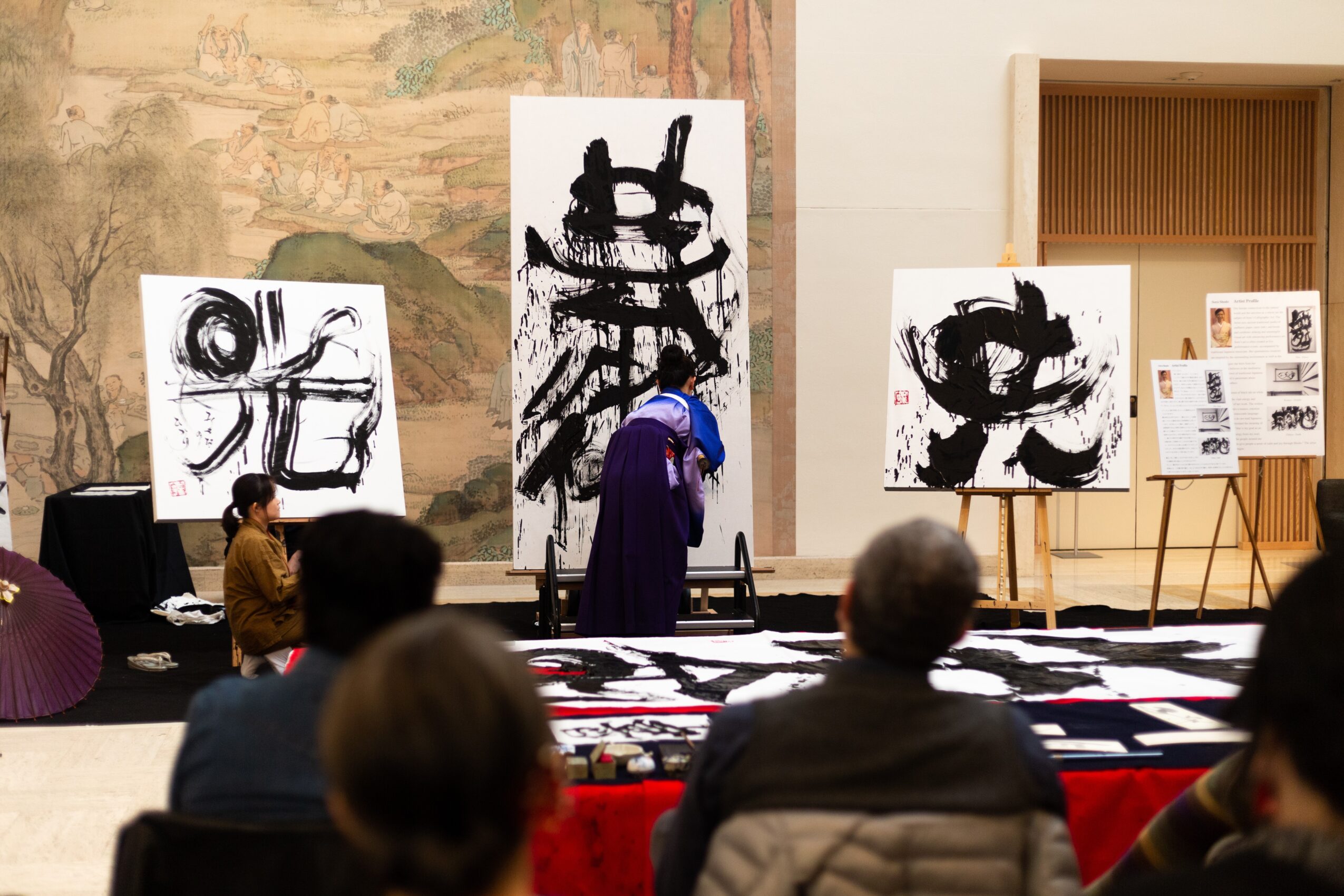 People observing large black and white calligraphy artworks displayed on easels and tables in an indoor gallery space.