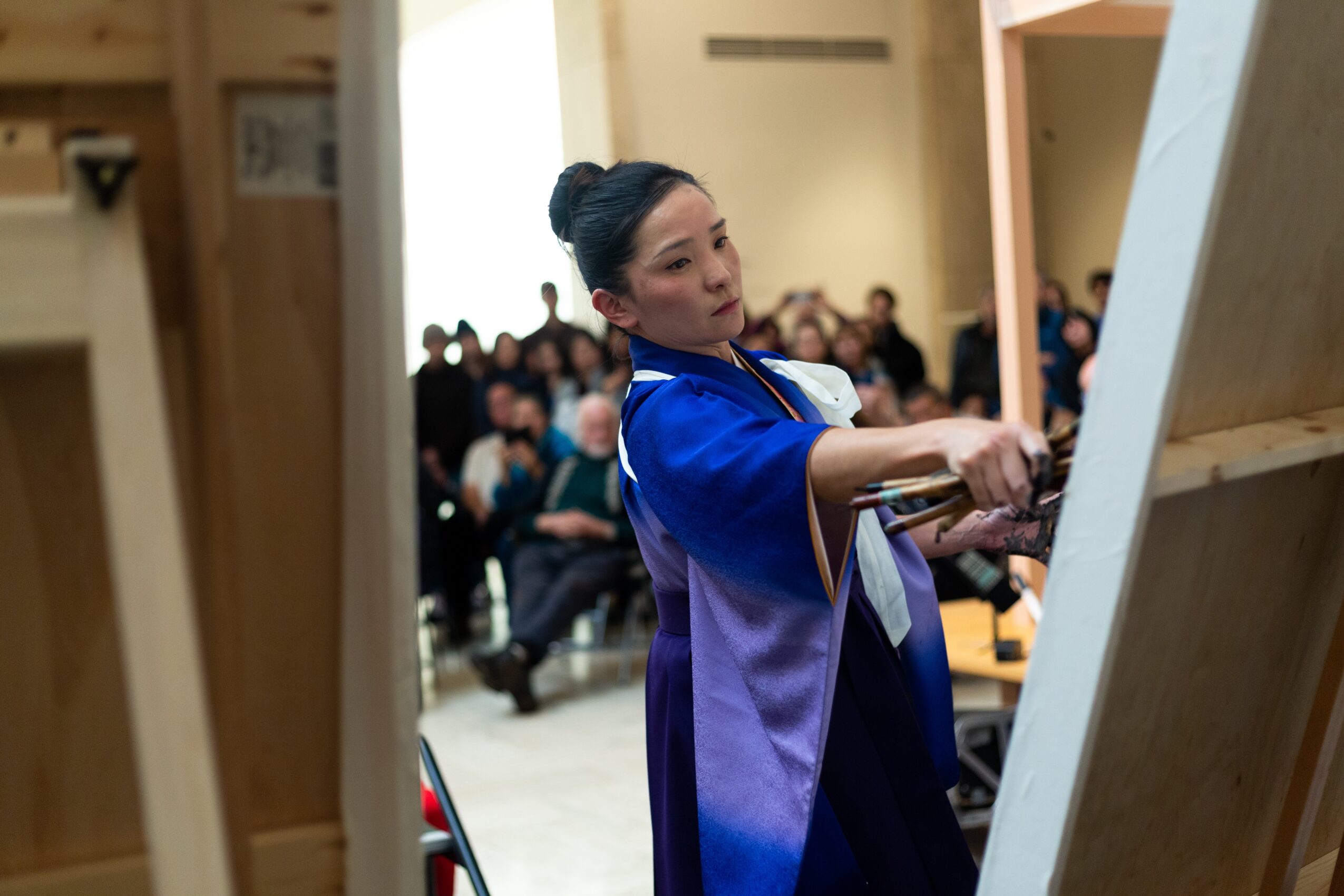 A woman in a blue and purple traditional dress demonstrates calligraphy to an audience in a room with wooden walls and seated spectators.