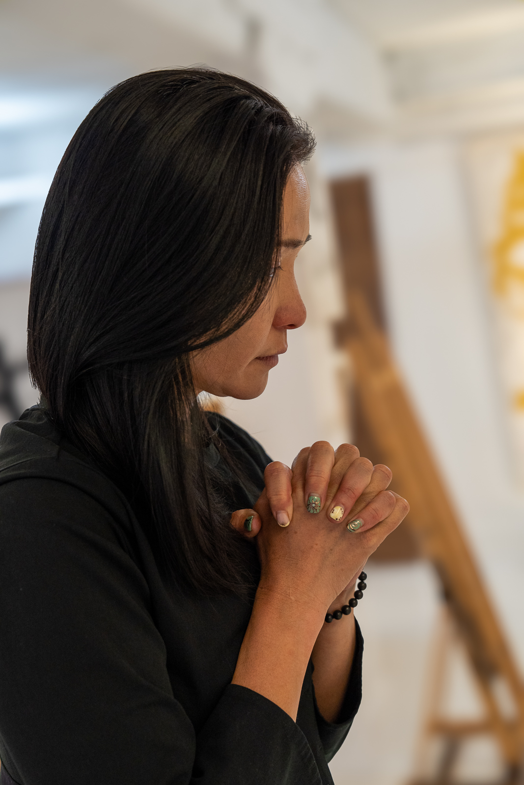 Woman with dark hair and closed eyes praying with hands clasped, indoors, neutral background.