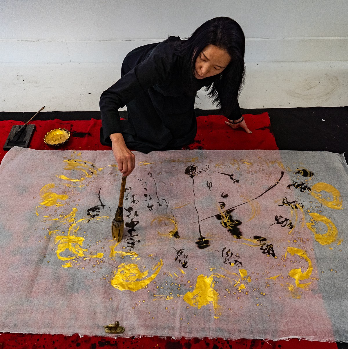 Woman painting calligraphy on fabric, kneeling on floor, with red and black mats nearby.