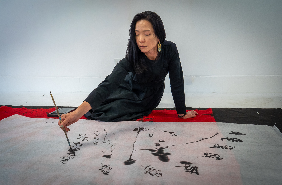 Woman sitting on floor, painting calligraphy on large paper, with a focused expression, against a plain background.