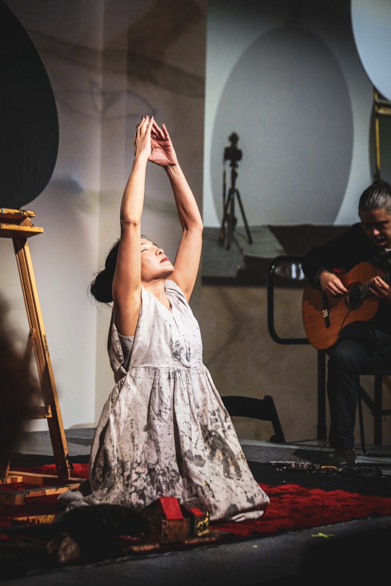 A woman kneels on a stage with her arms raised, eyes closed, and a man plays guitar beside her.