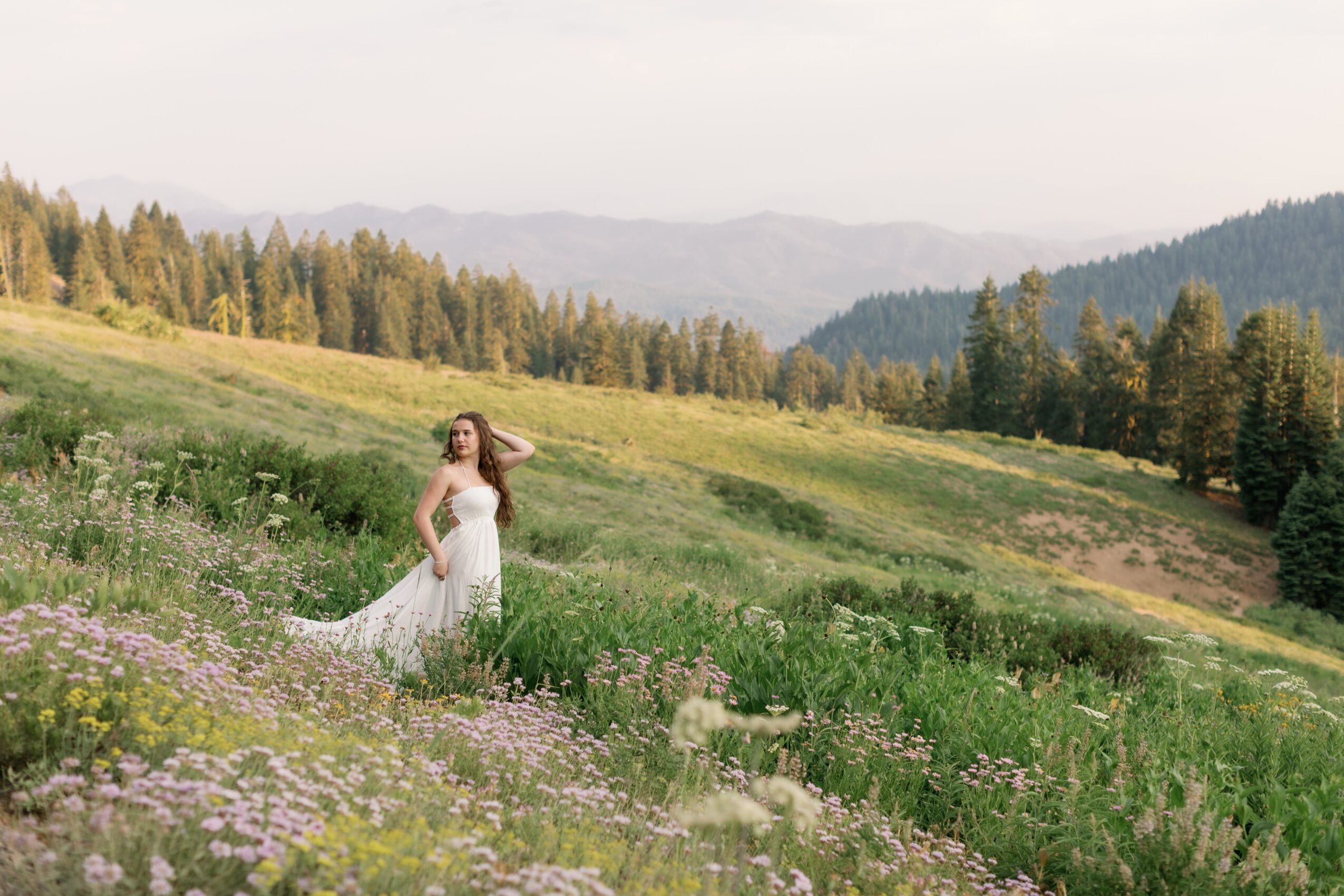 Woman in a white dress standing in a green meadow with rolling hills and trees in the background.