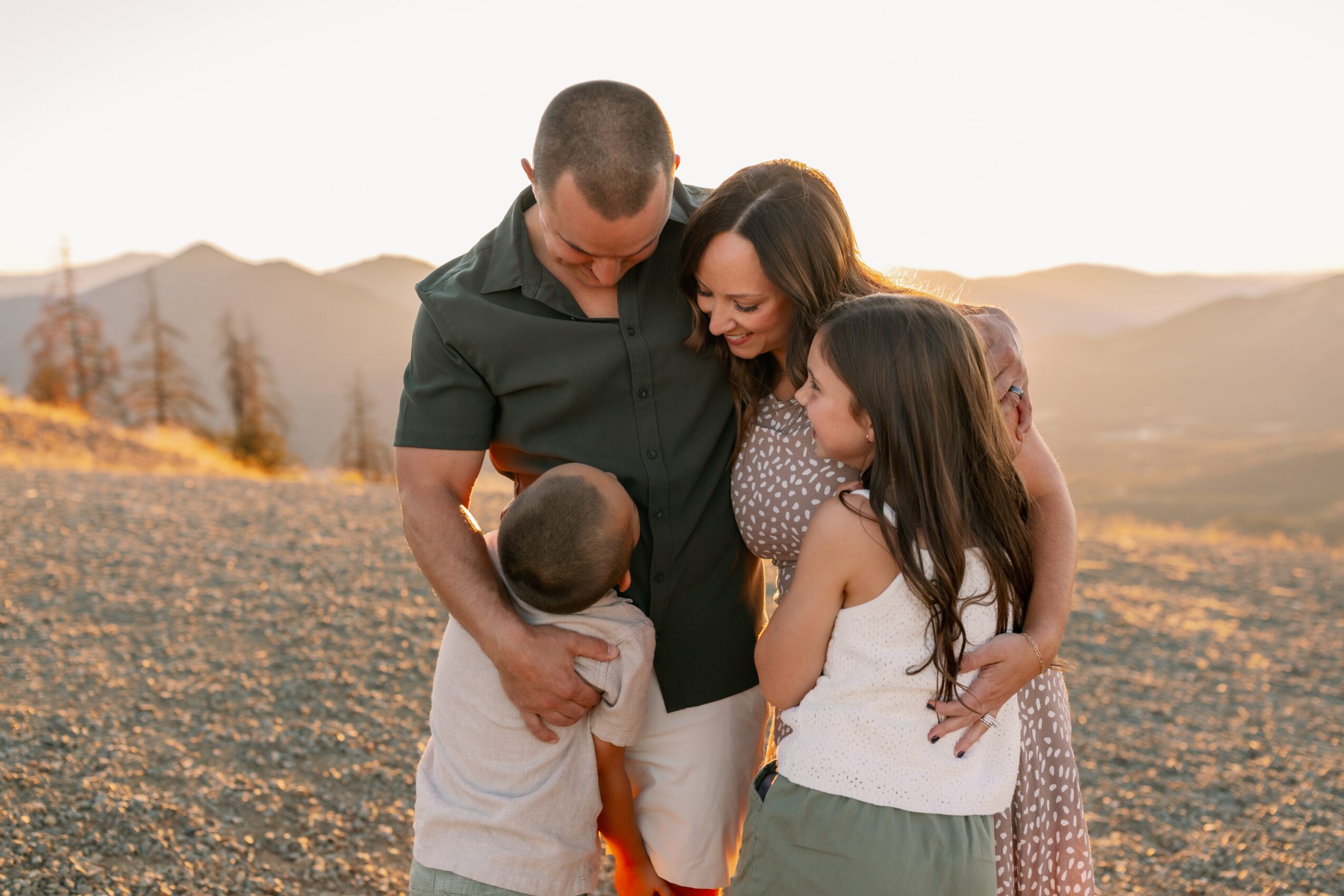 Family of five hugging outdoors with mountains in the background, sunset lighting, and smiling faces.