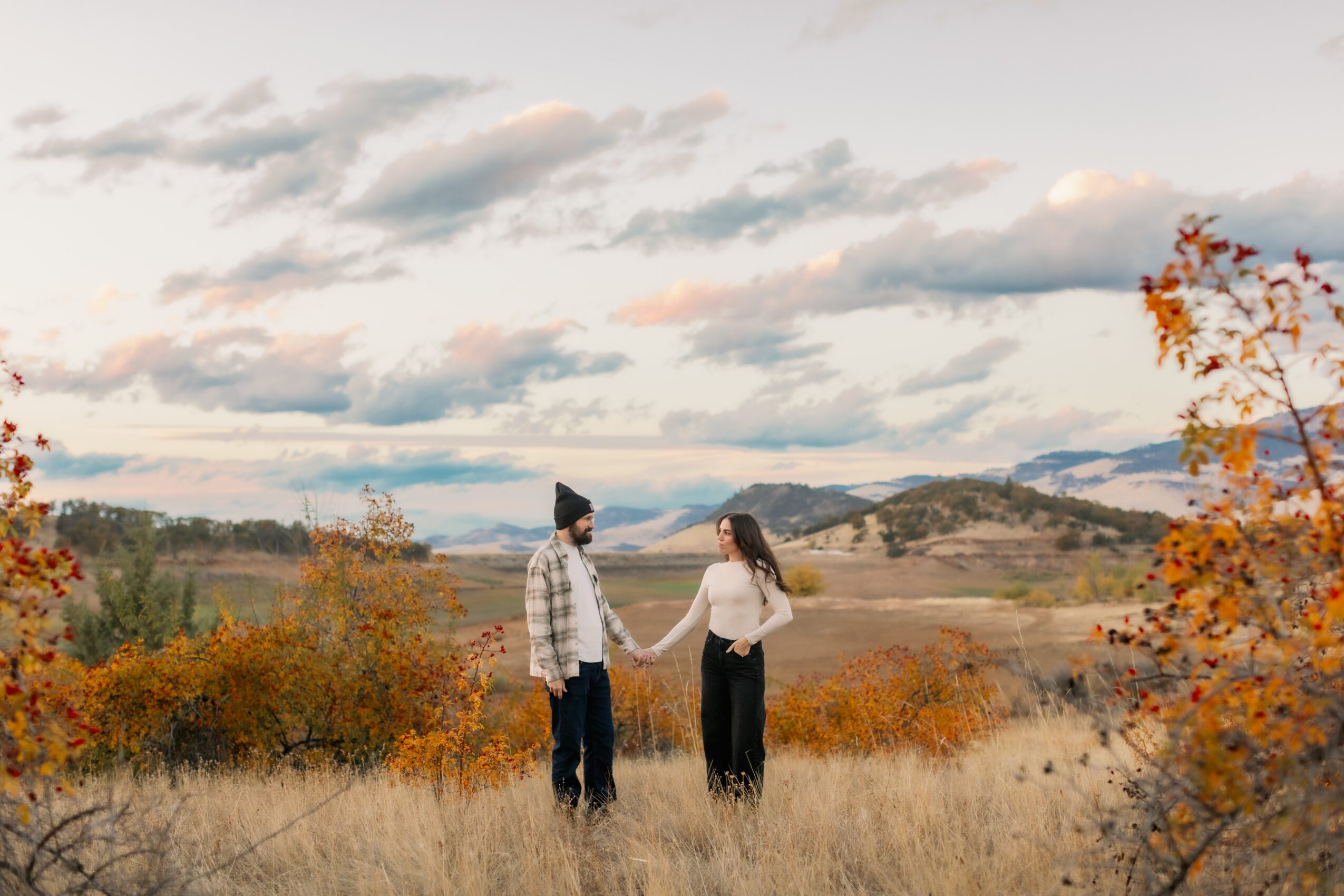 Two people holding hands outdoors with a landscape of hills, trees, and a cloudy sky in the background.