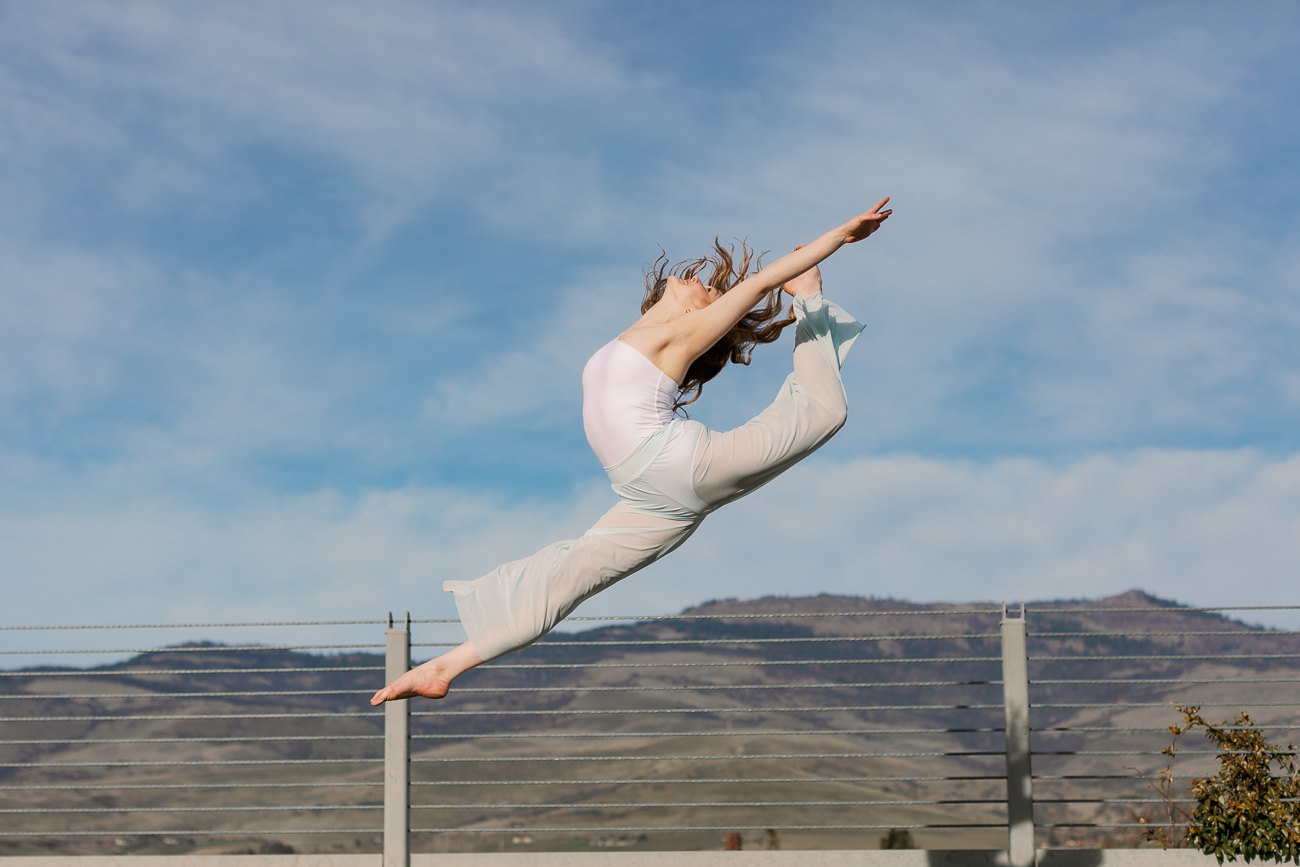Person jumping in the air outdoors with a blue sky and clouds, arms and legs extended, over a landscape with terraces.