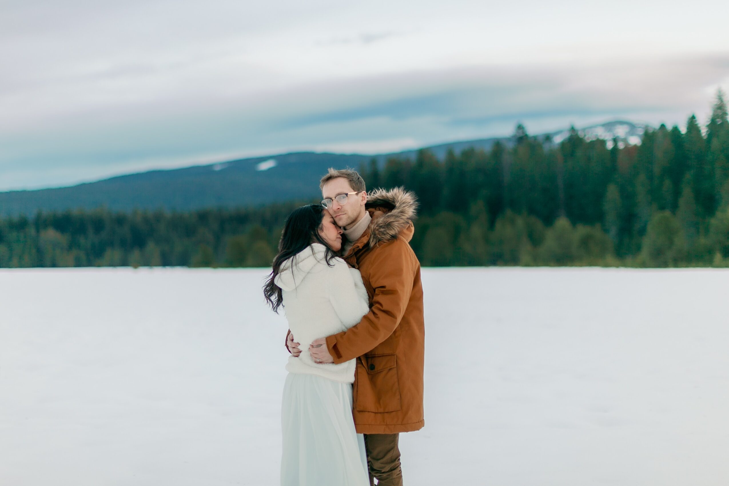 Couple embracing outdoors near a frozen lake with forest and cloudy sky in background.