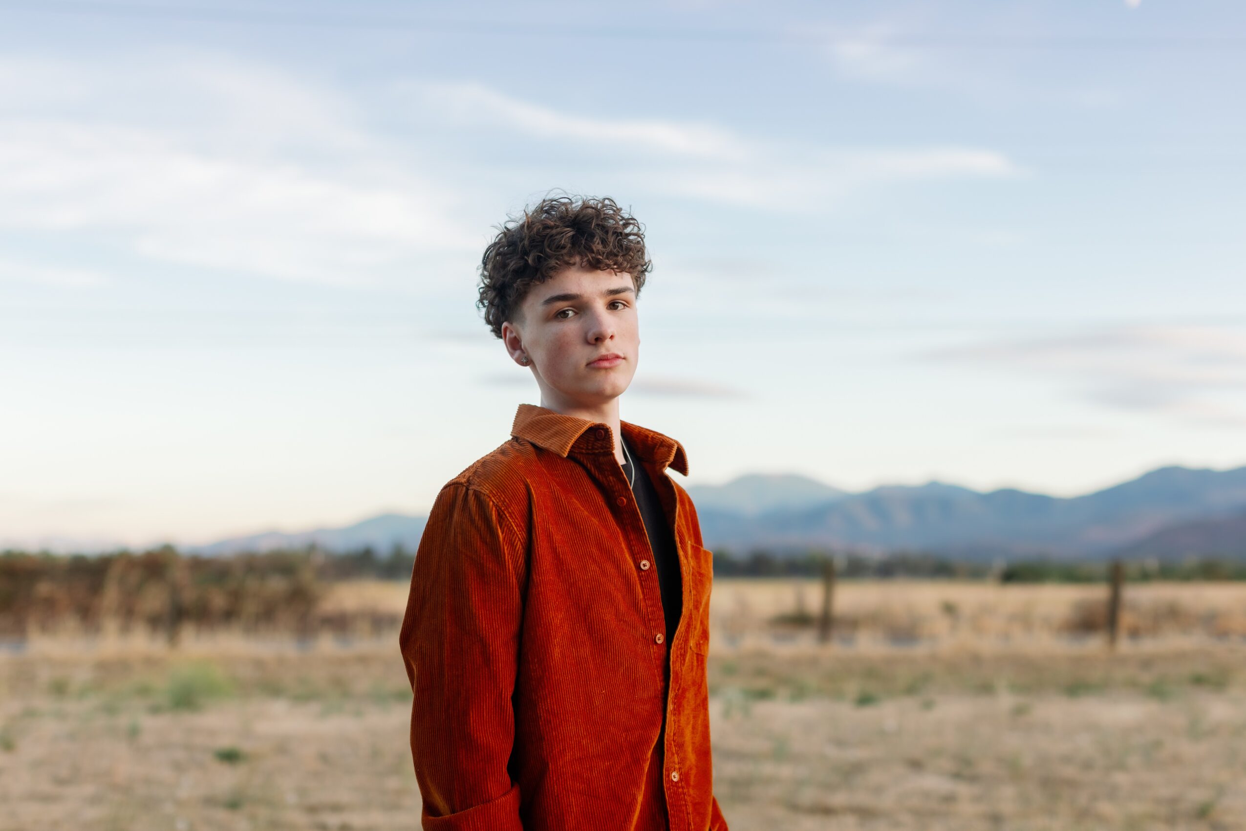 Young person with curly hair standing outdoors in a field with mountains in the background.