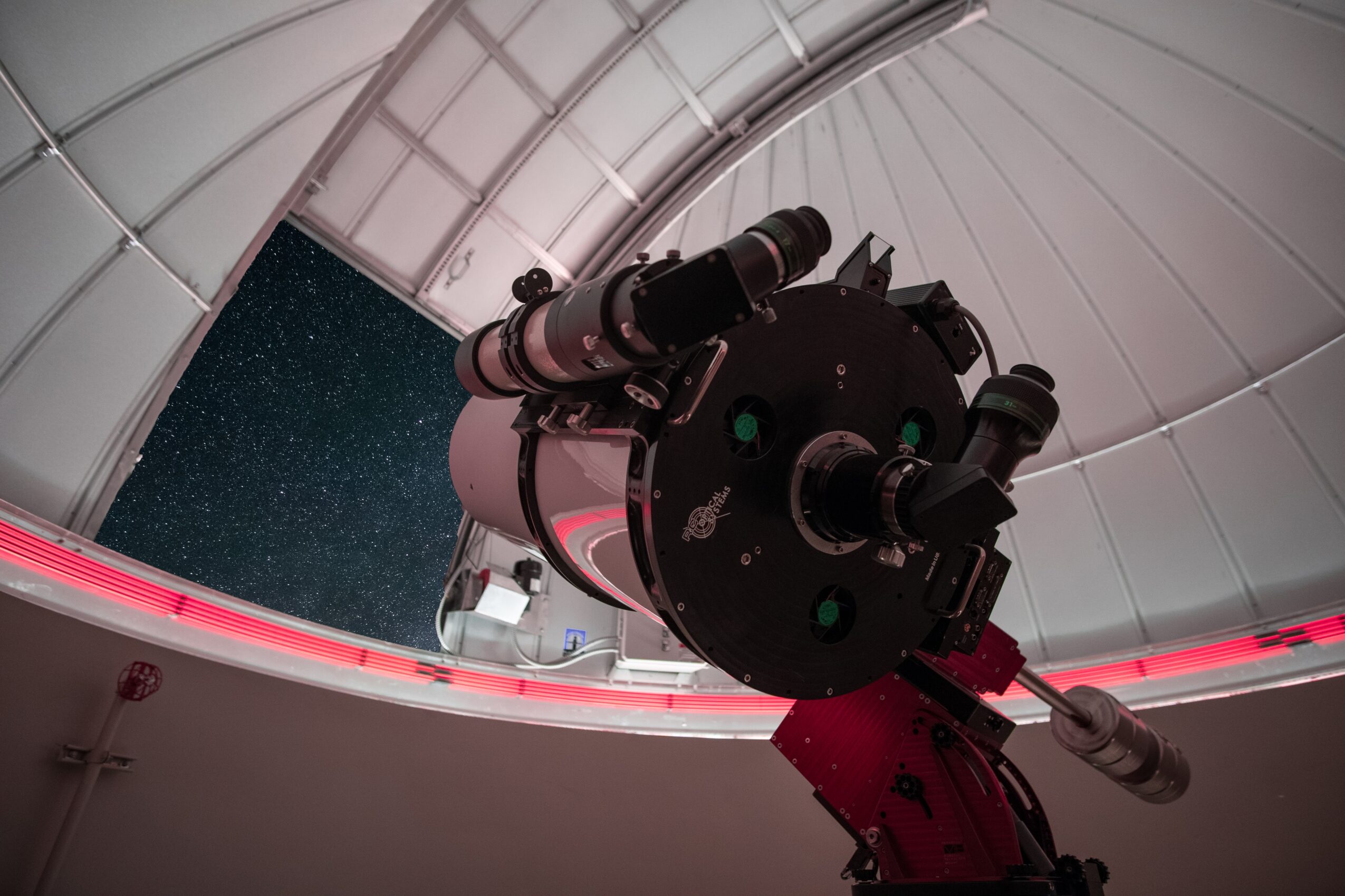Telescope inside an observatory dome, pointing upward at night sky with stars visible through the opening.