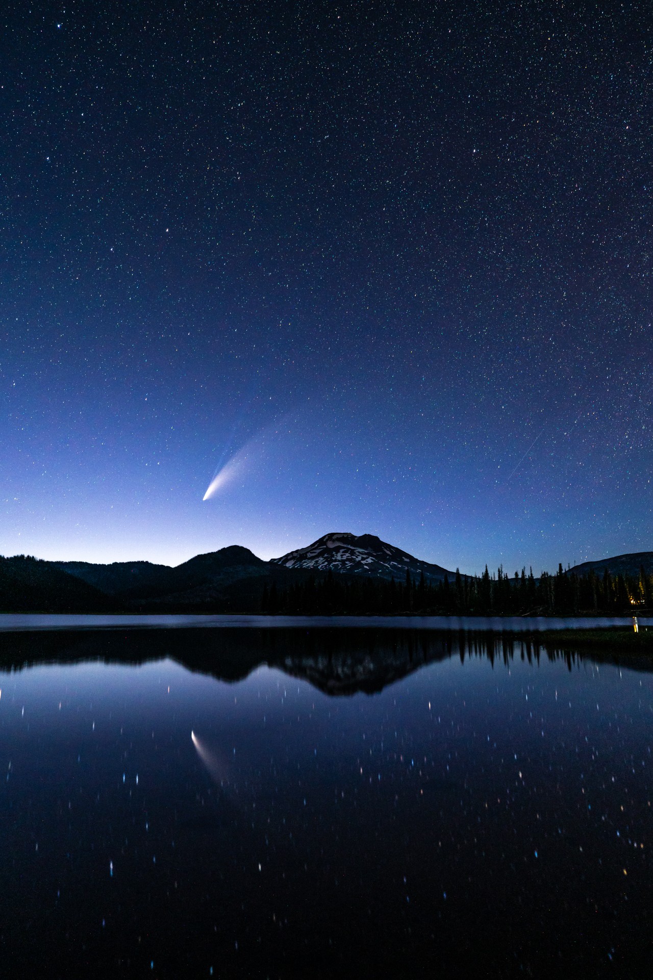 Night sky with stars and a comet above mountains reflected in a lake.