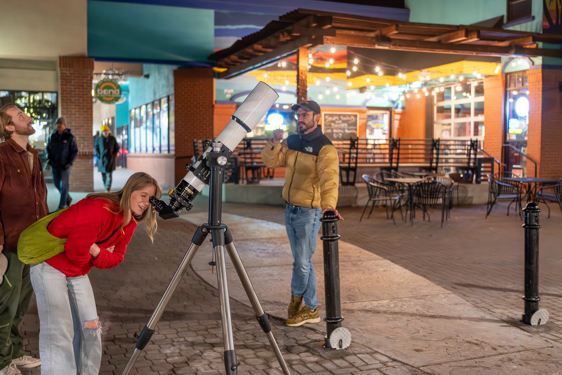Two people, a woman in a red jacket and a man in a yellow jacket, look through a telescope outside a building with colorful lights.