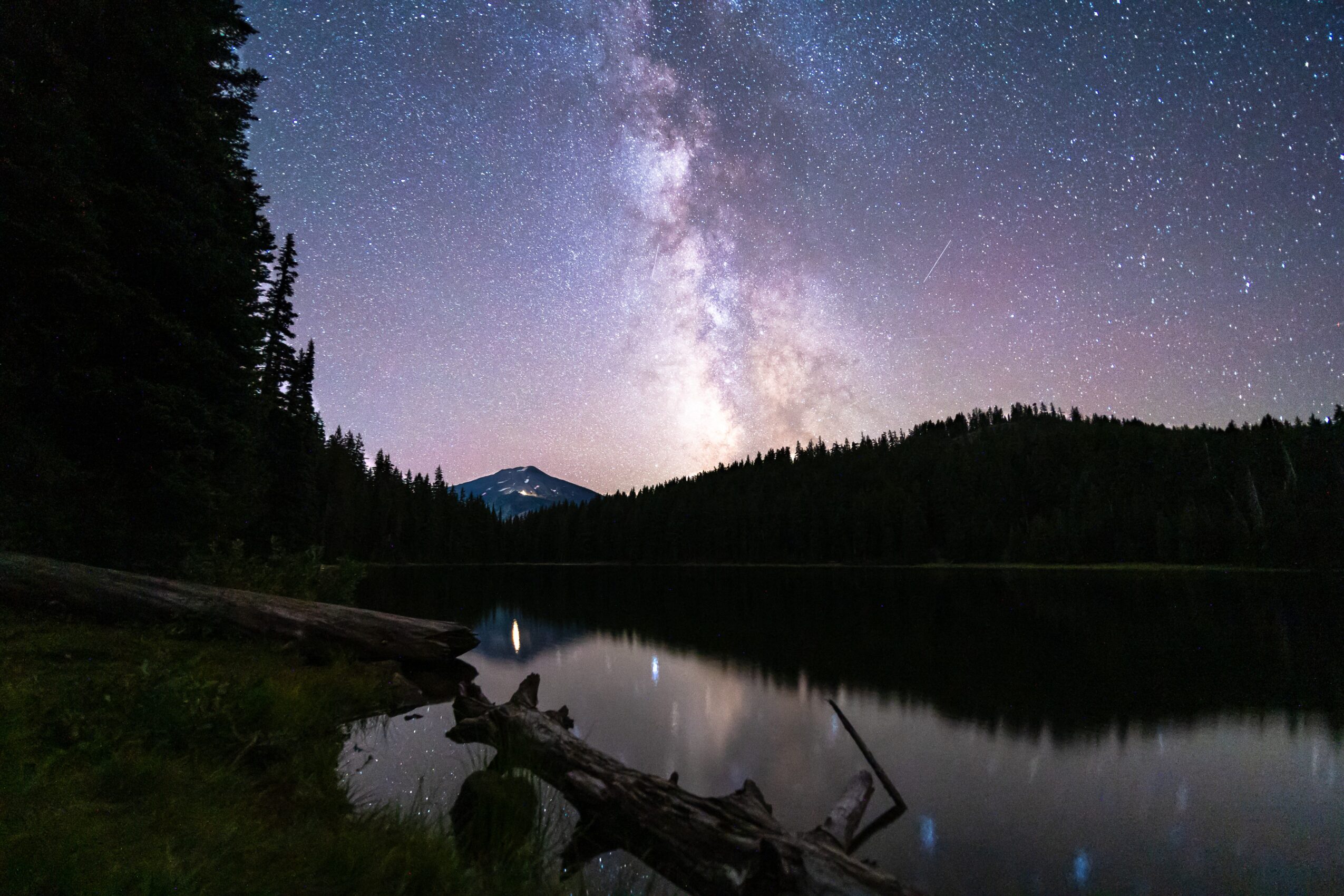 Night sky with stars and the Milky Way above a calm lake, surrounded by trees and mountains.