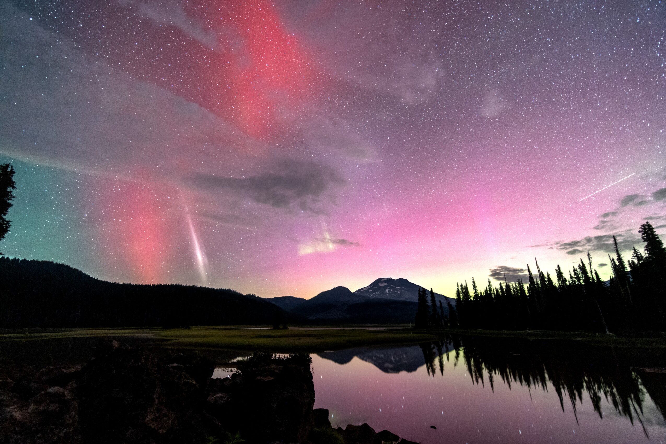 Night sky with stars, colorful aurora, and a mountain silhouette reflected in a lake, with trees on the right side.