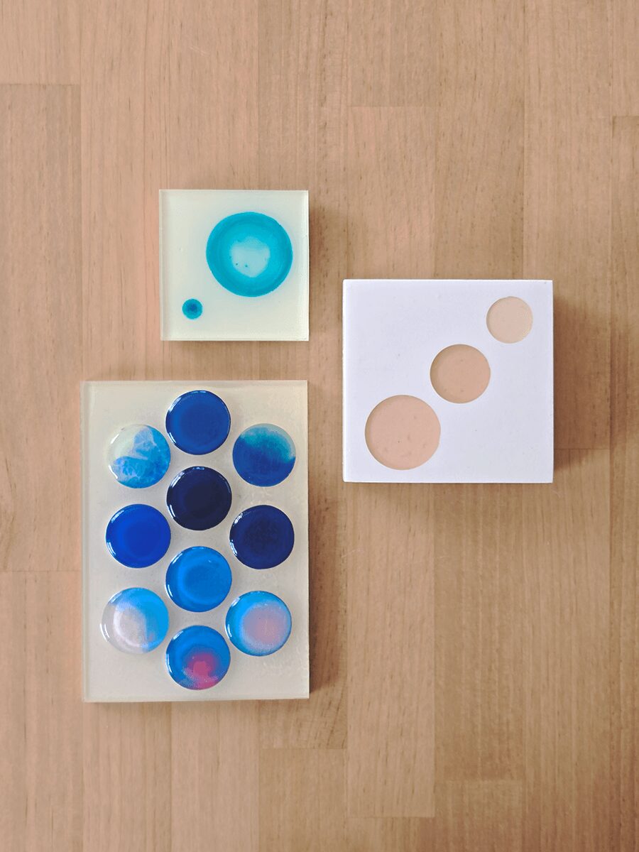 Three white square trays with colorful circular objects on a wooden surface.