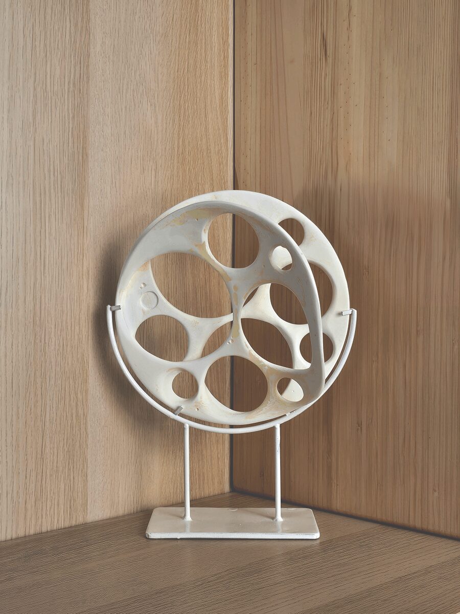 White circular sculpture with various holes, mounted on a stand against a wooden wall.