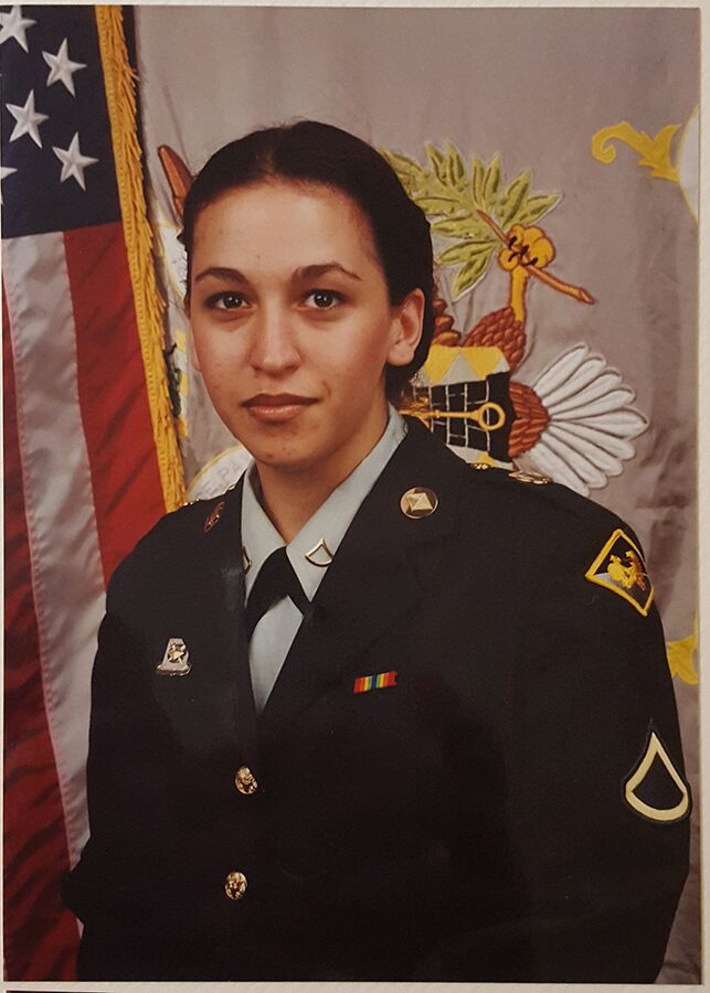 Young woman in military uniform standing in front of flags and emblem, looking at camera, neutral expression.