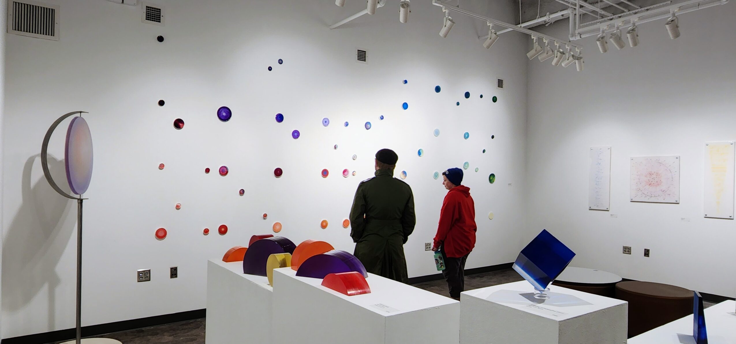 Two people stand in an art gallery with colorful dots on the wall, and display tables with objects, in a well-lit room.