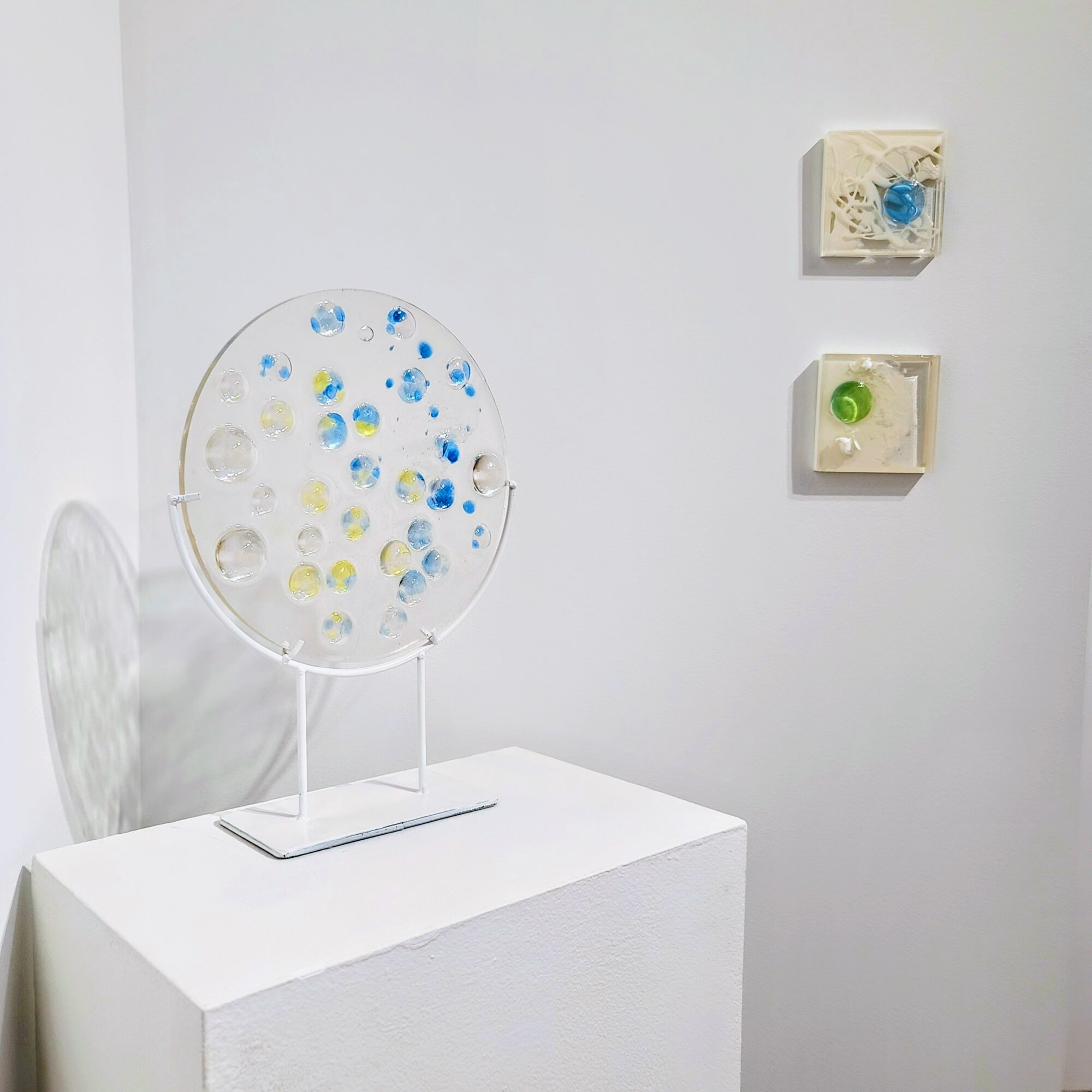 White display stand with a circular art piece featuring blue, yellow, and clear elements, and two small wall-mounted boxes with objects inside.