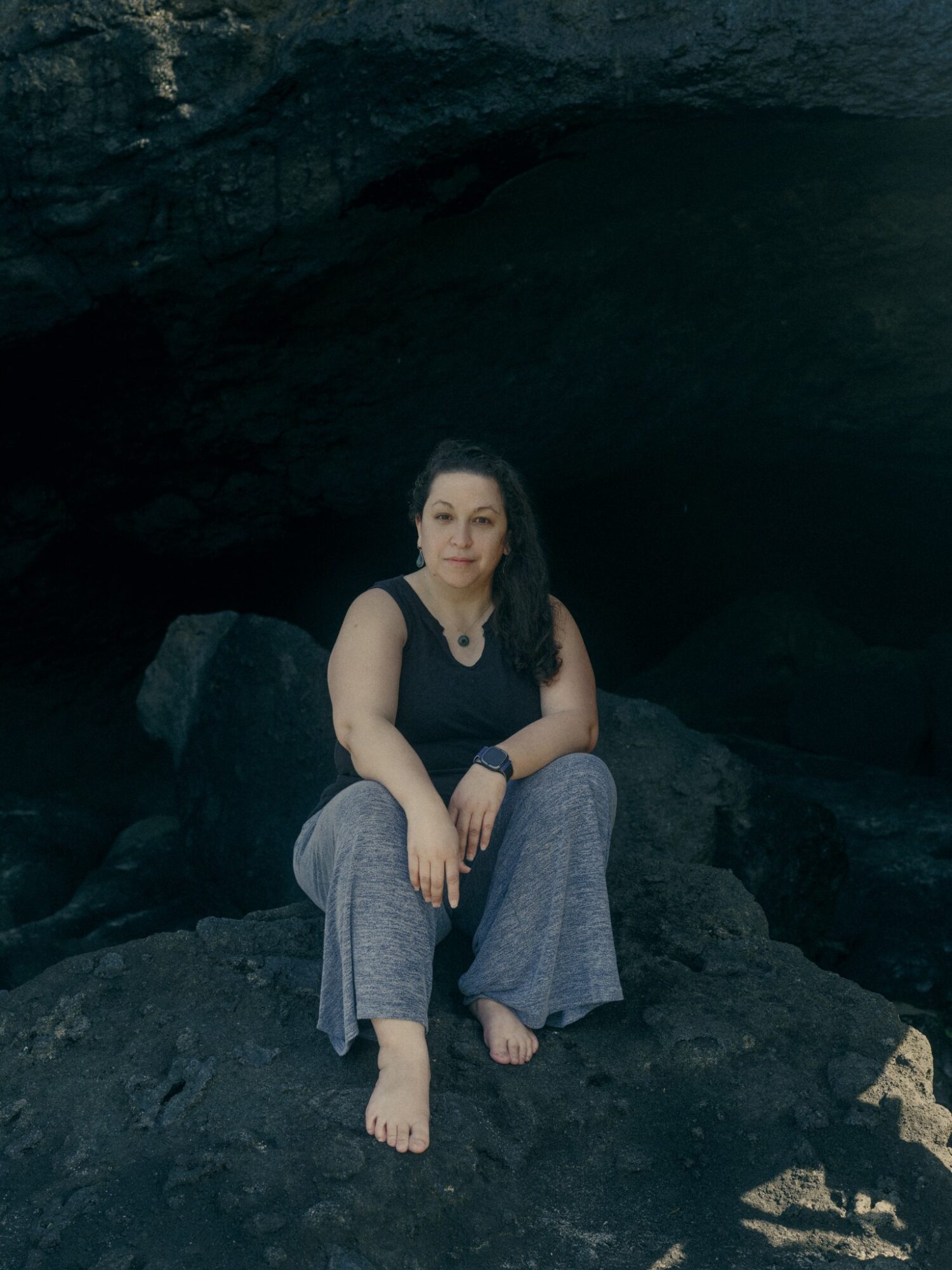 Woman sitting on rocks in a dark cave, wearing a black top and gray pants, with long dark hair.