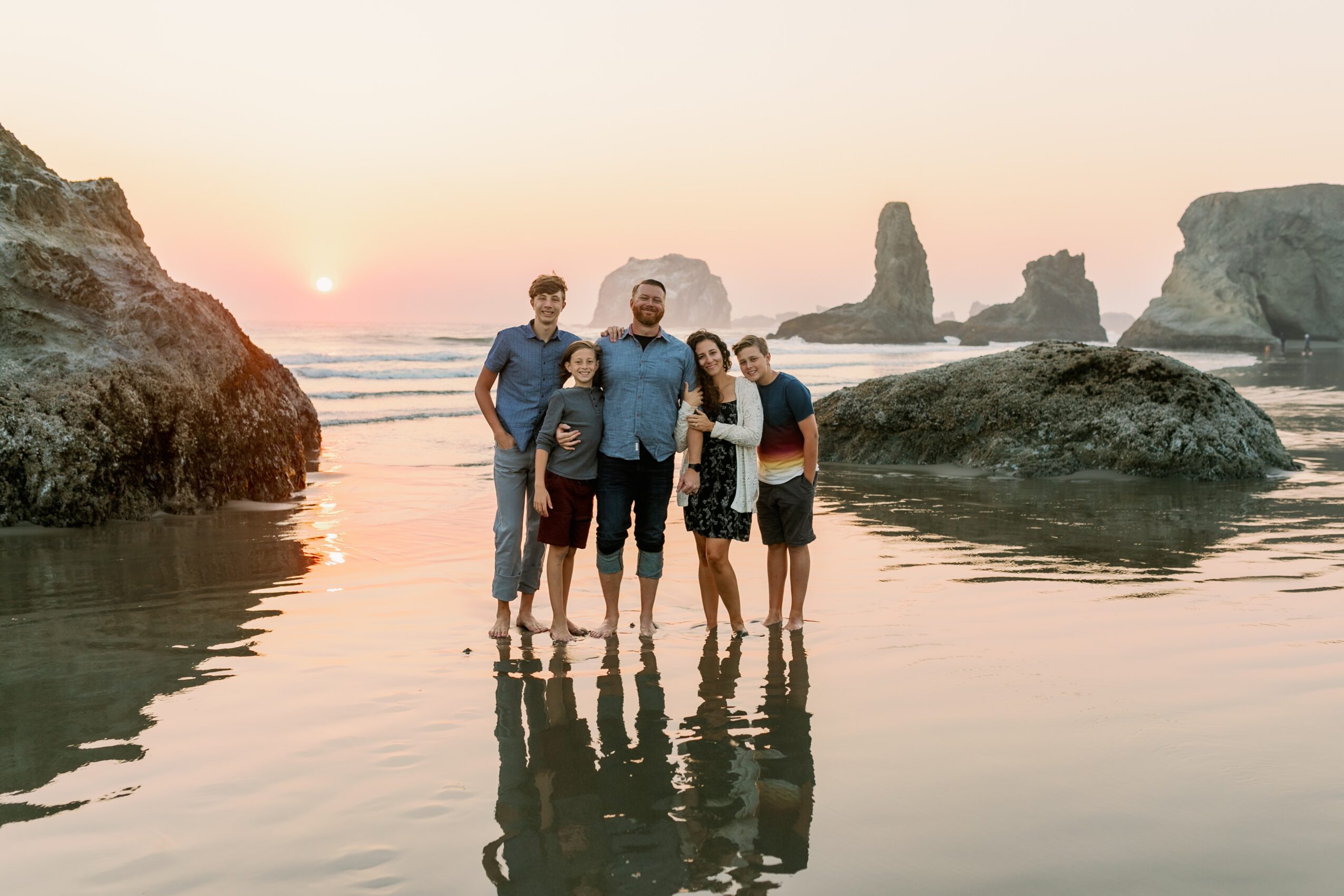 Group of five people standing in shallow water at the beach during sunset, with rocks and sea stacks in background.