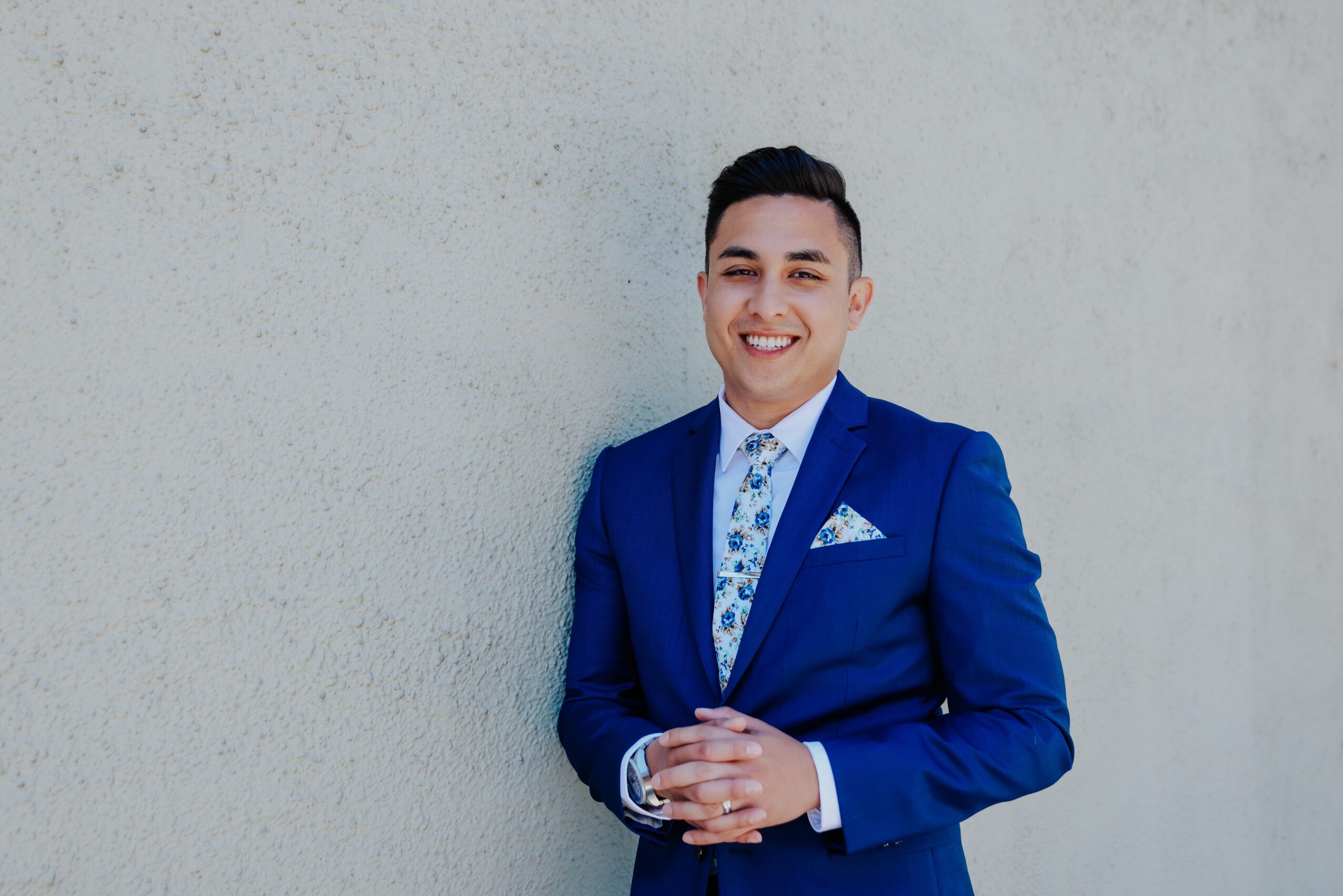 Young man in a blue suit smiling, standing against a plain wall, hands clasped, wearing a patterned shirt and pocket square.