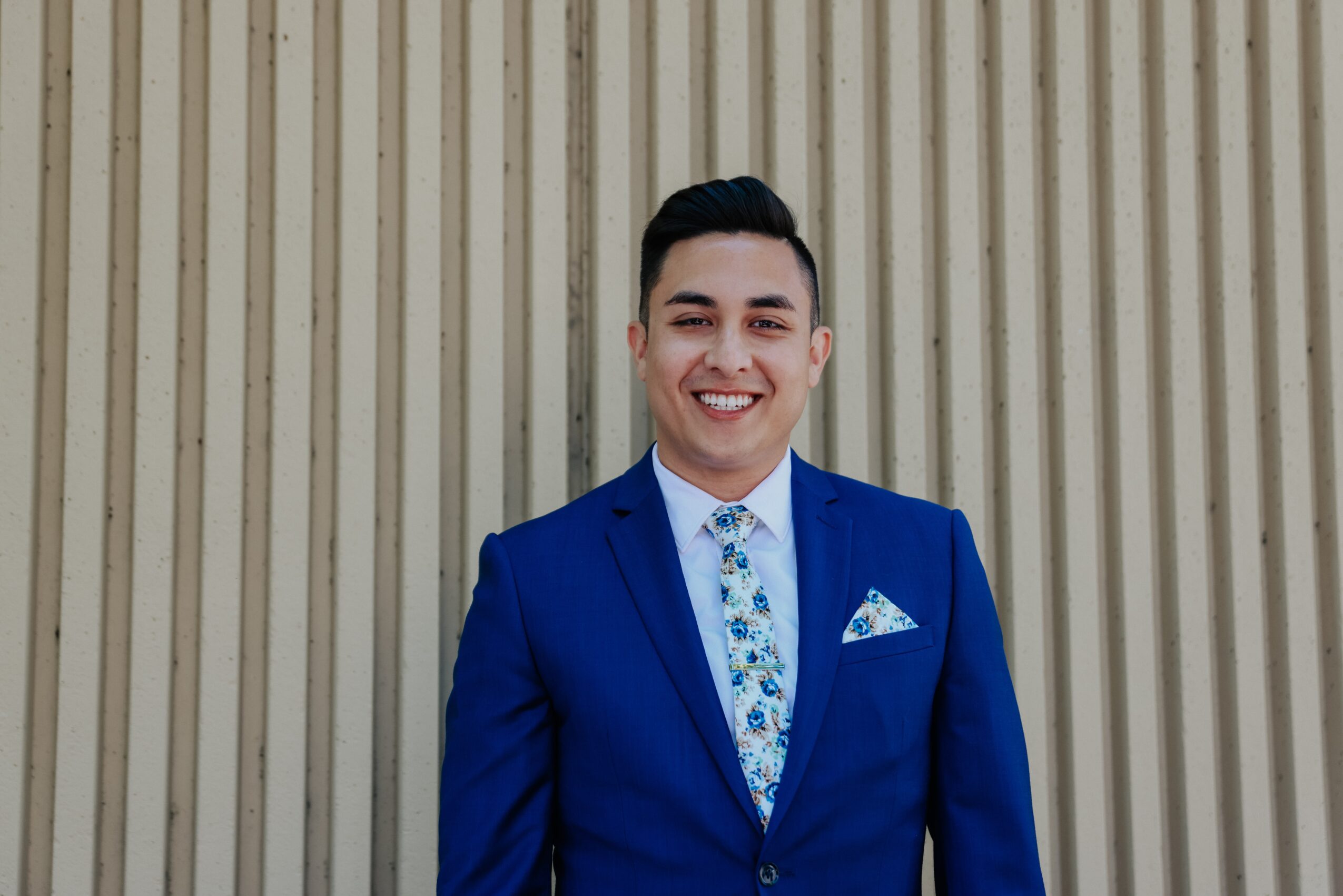 Young man in a blue suit smiling, standing in front of a beige corrugated wall.