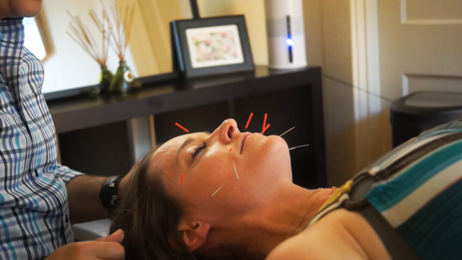 Woman lying down with eyes closed, receiving a facial treatment in a room with framed pictures and decor.