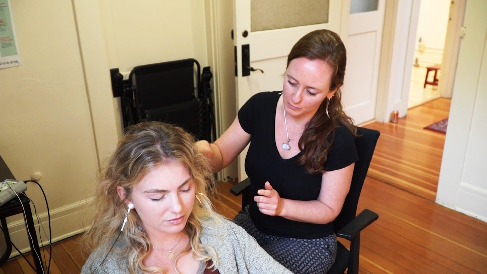 Two women in a room, one sitting with eyes closed, the other standing and touching her shoulder, in a home setting.