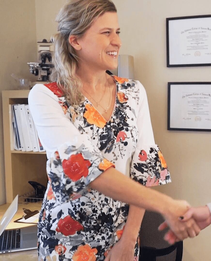 Smiling woman in floral dress shaking hands in an office with certificates on the wall.