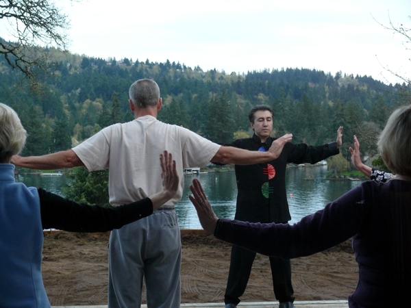 Four people stand outdoors near a lake with trees in the background, holding their arms out in a circle.