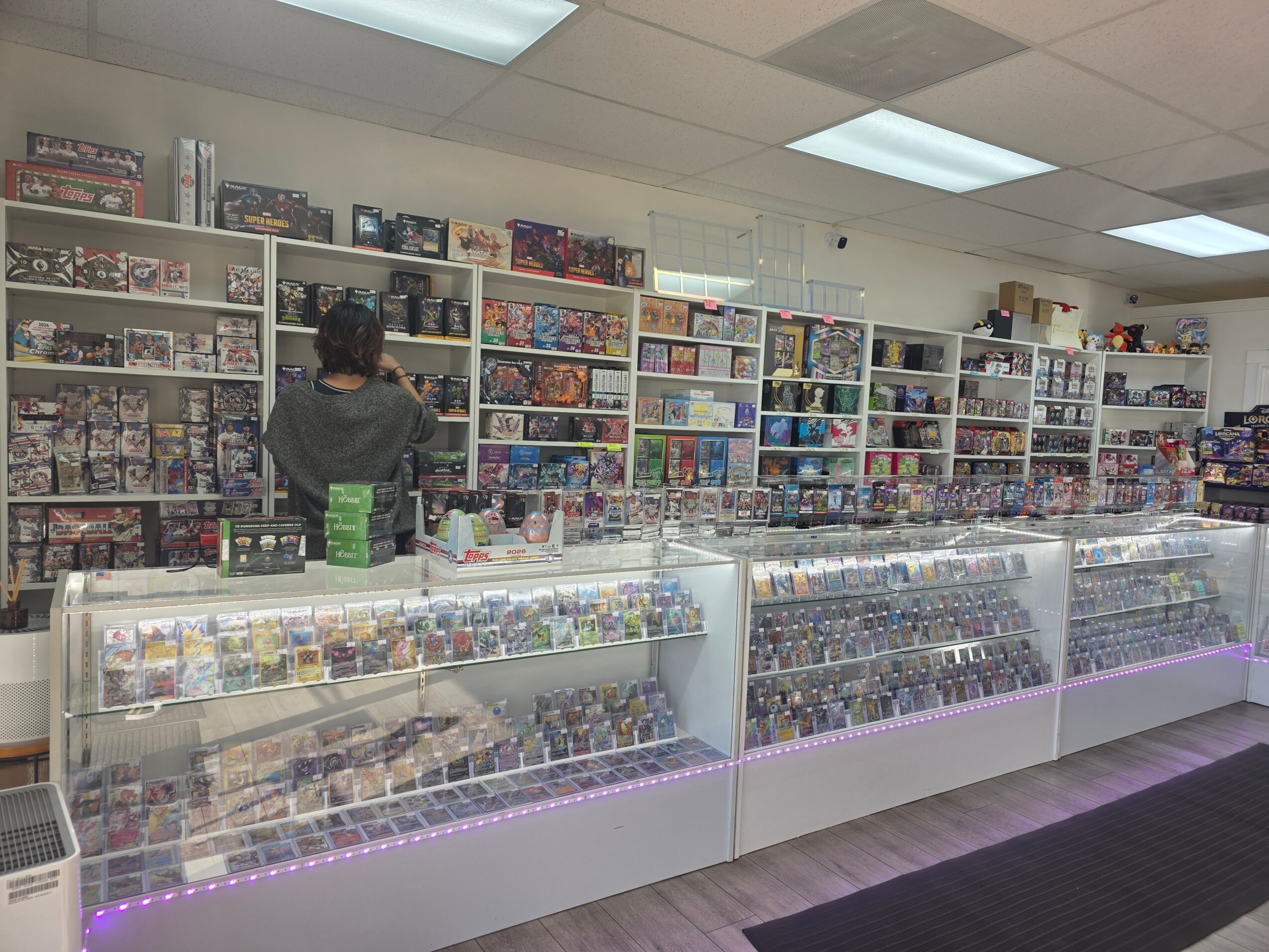 Person browsing shelves filled with board games and collectibles in a store, with glass display cases in front, ceiling lights, and a black floor mat.