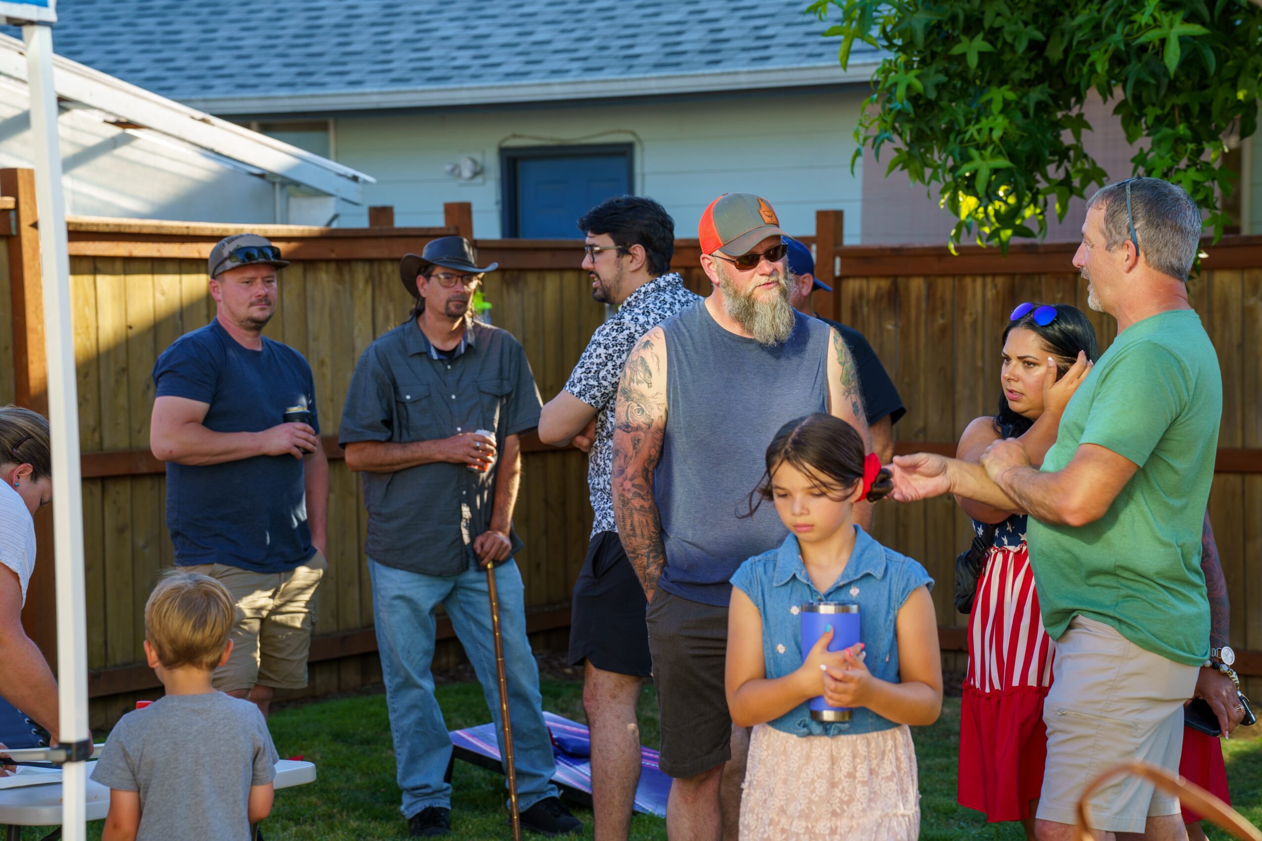 Group of people talking outdoors in a backyard, with children nearby, wooden fence, house, and trees in background.