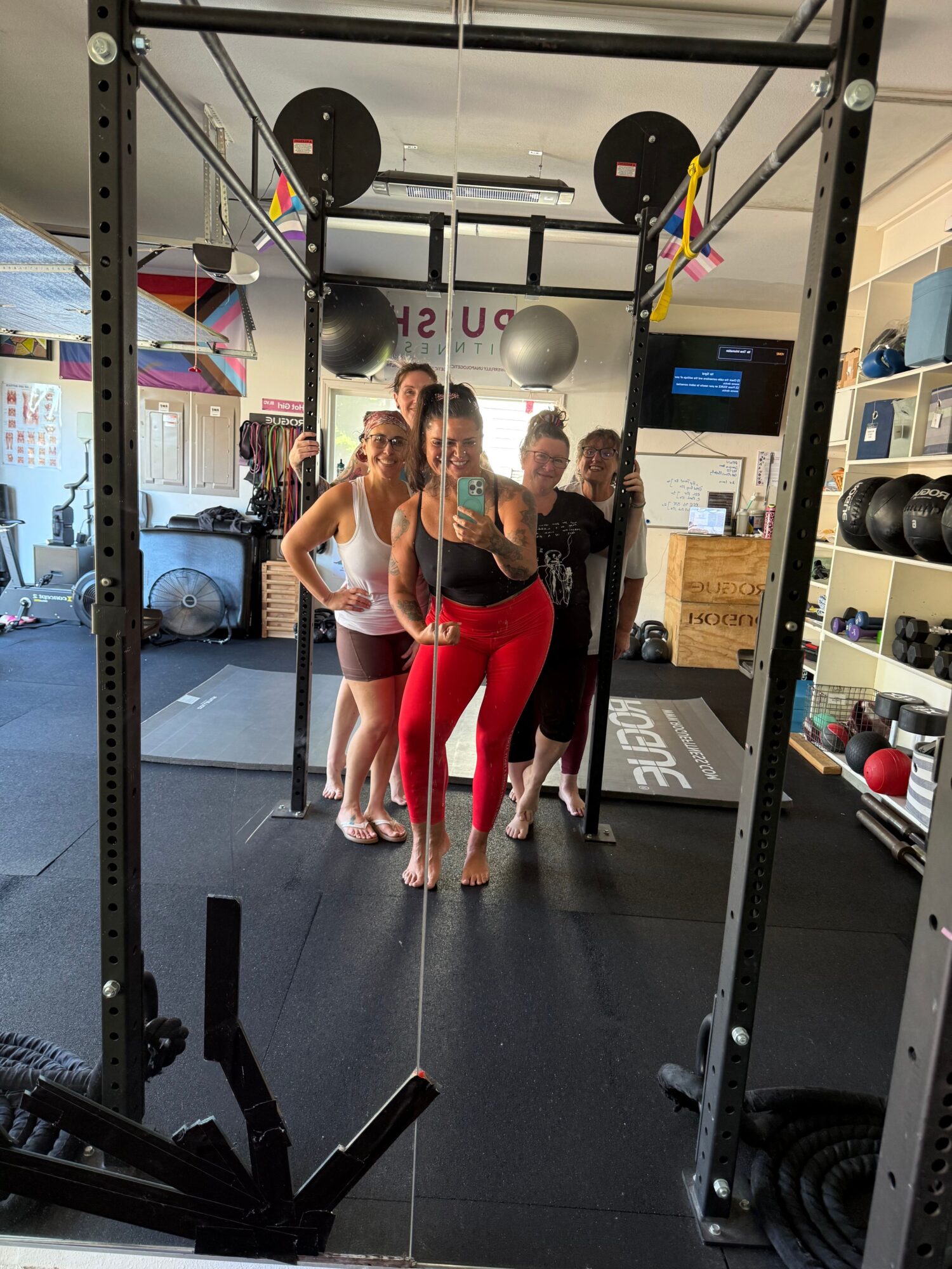 People in a gym taking a mirror selfie, with gym equipment and weights visible in the background.