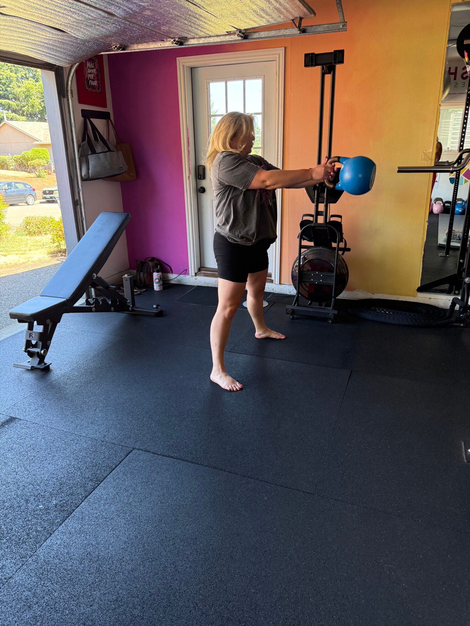 Person with blonde hair in a gym holding a blue kettlebell, standing on a black floor with gym equipment around.