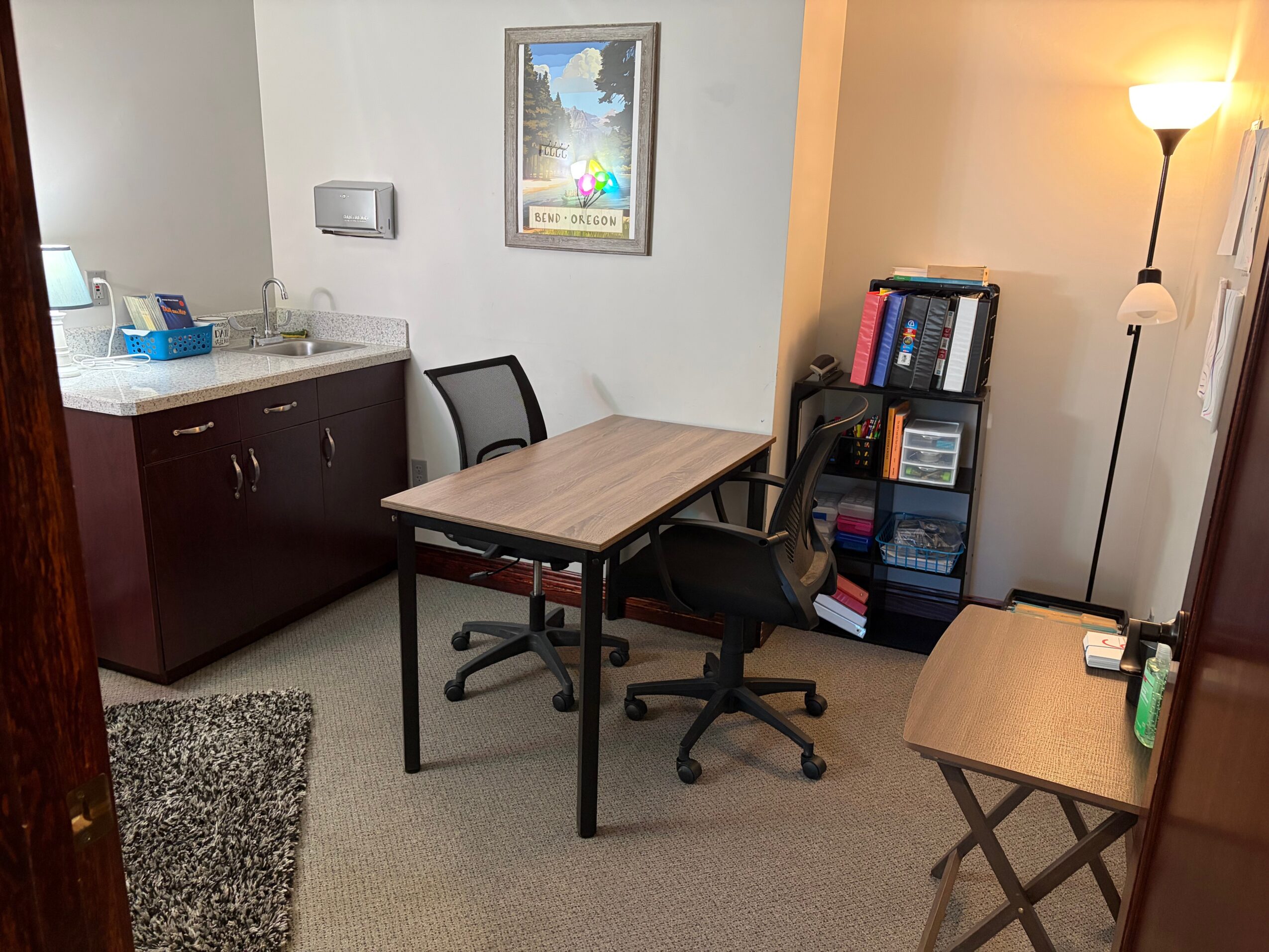 Office corner with desk, two chairs, bookshelf, and a floor lamp, with a framed picture on the wall.