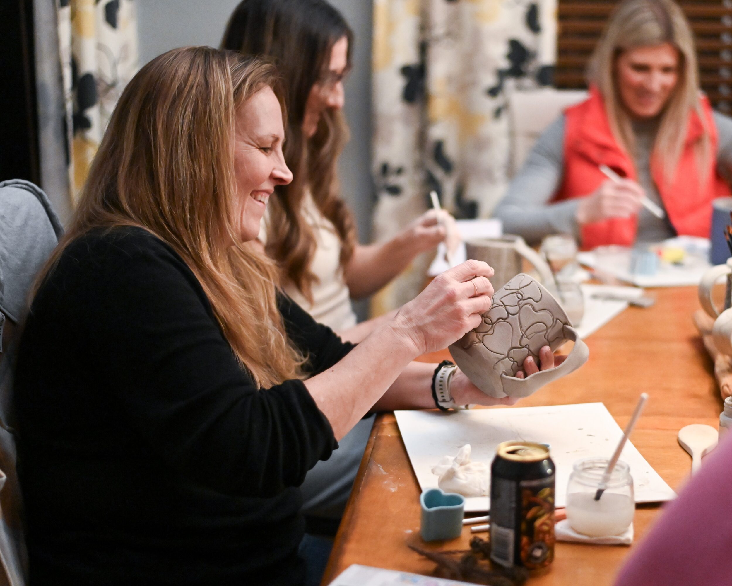 Three women sit at a table, one painting a ceramic object, with art supplies and drinks on the table.