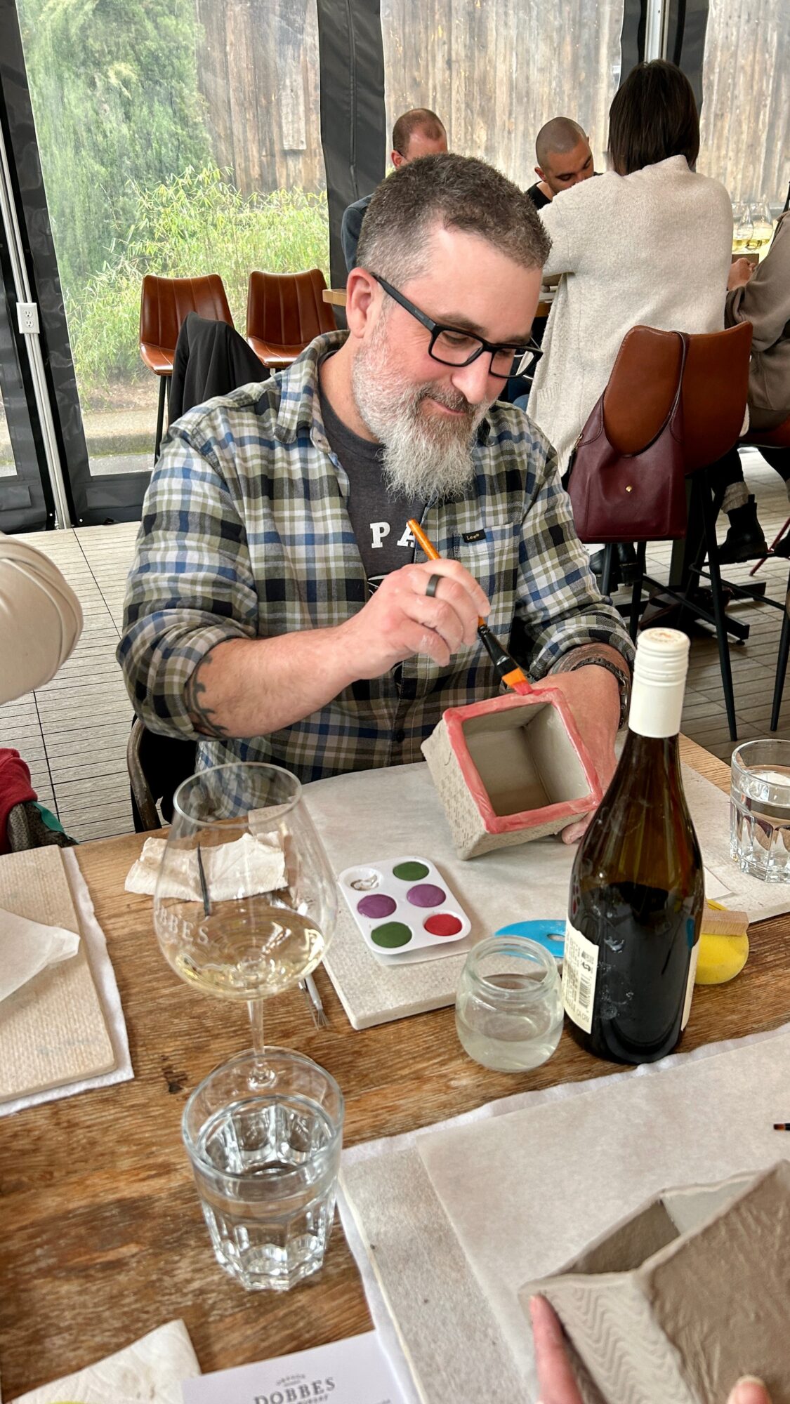 Man with glasses and beard, wearing a plaid shirt, sitting at a table with makeup palette and drinks, smiling.