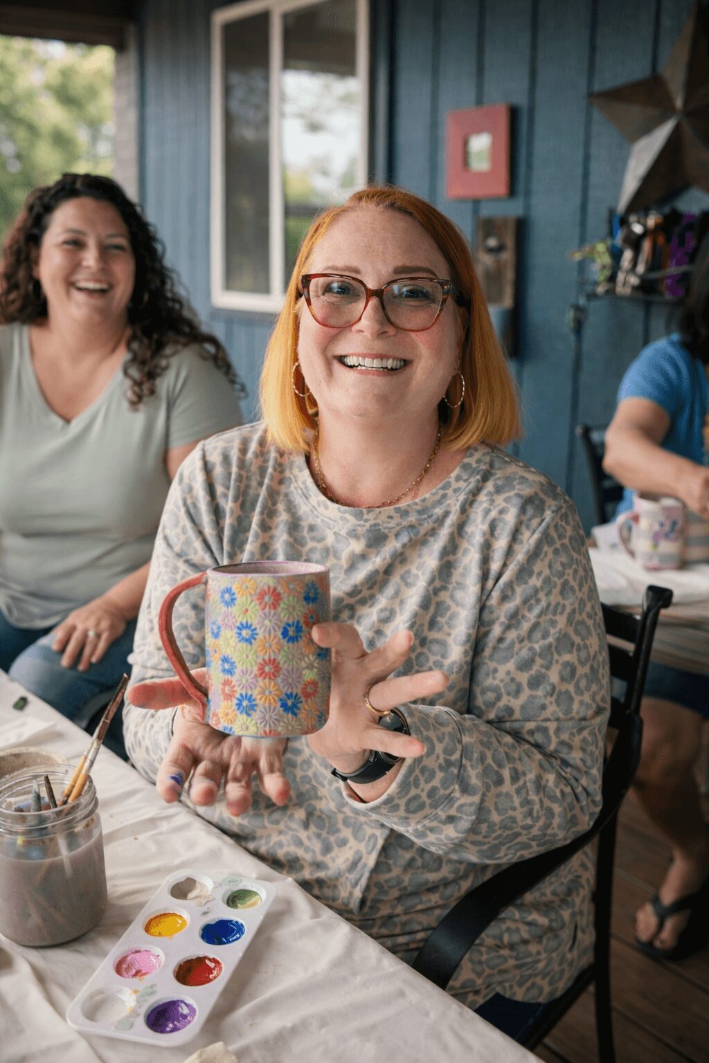 Two women smiling, one holding a colorful mug, sitting at a table with paint supplies, in a decorated room.
