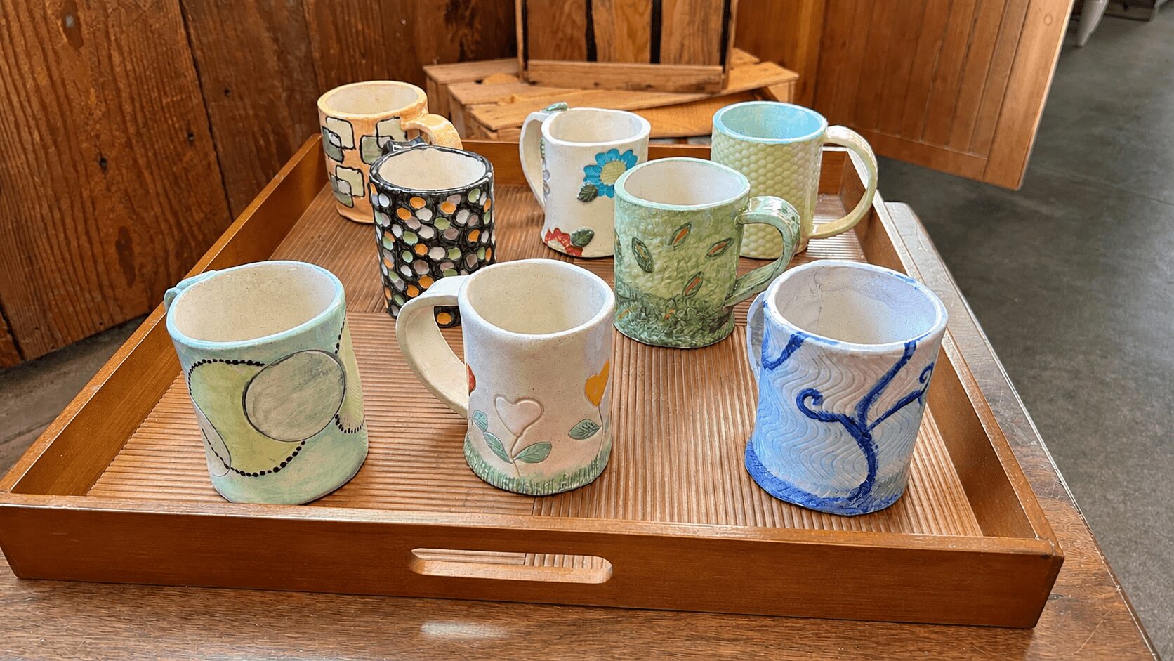 Collection of nine ceramic mugs with various colorful patterns on a wooden tray.