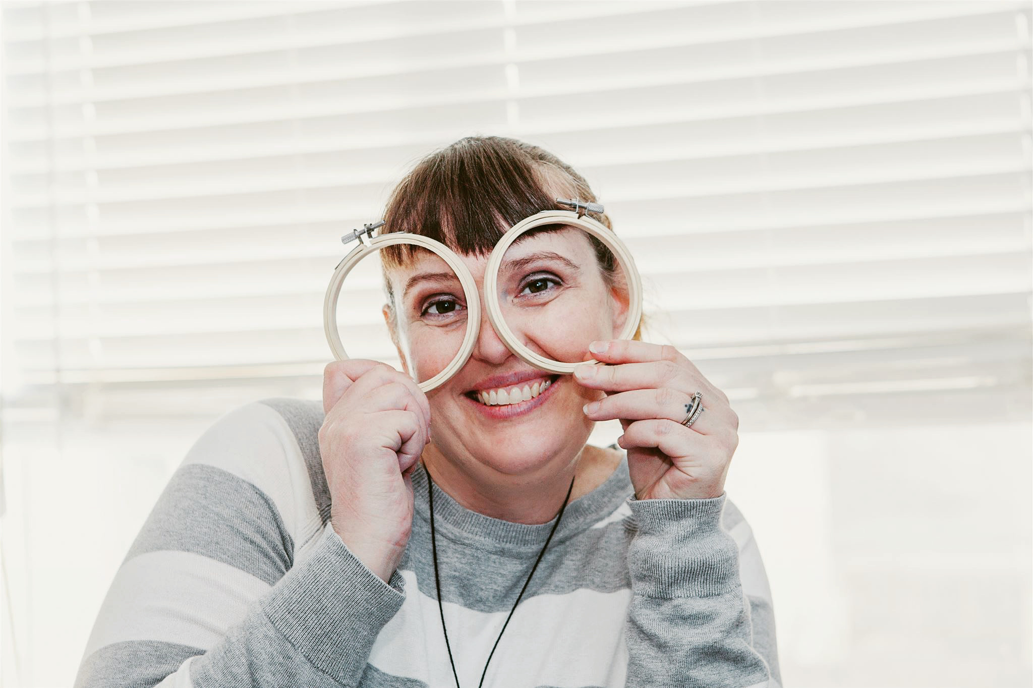 Person smiling and holding glasses in front of face, sitting near window with blinds, wearing a striped sweater.