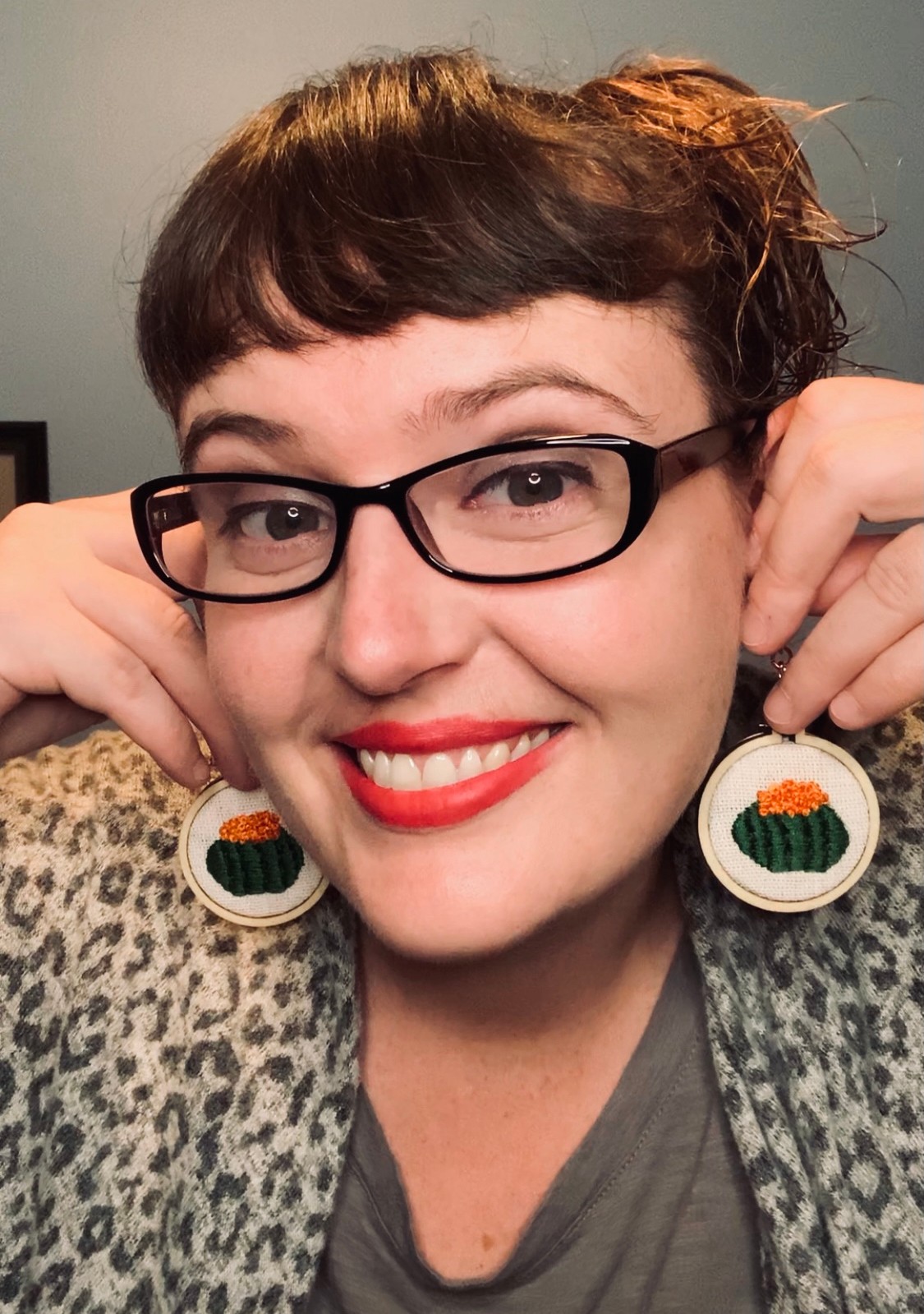 Woman with short dark hair, glasses, red lipstick, smiling, wearing earrings with pumpkin and striped pattern, gray top, leopard print jacket.