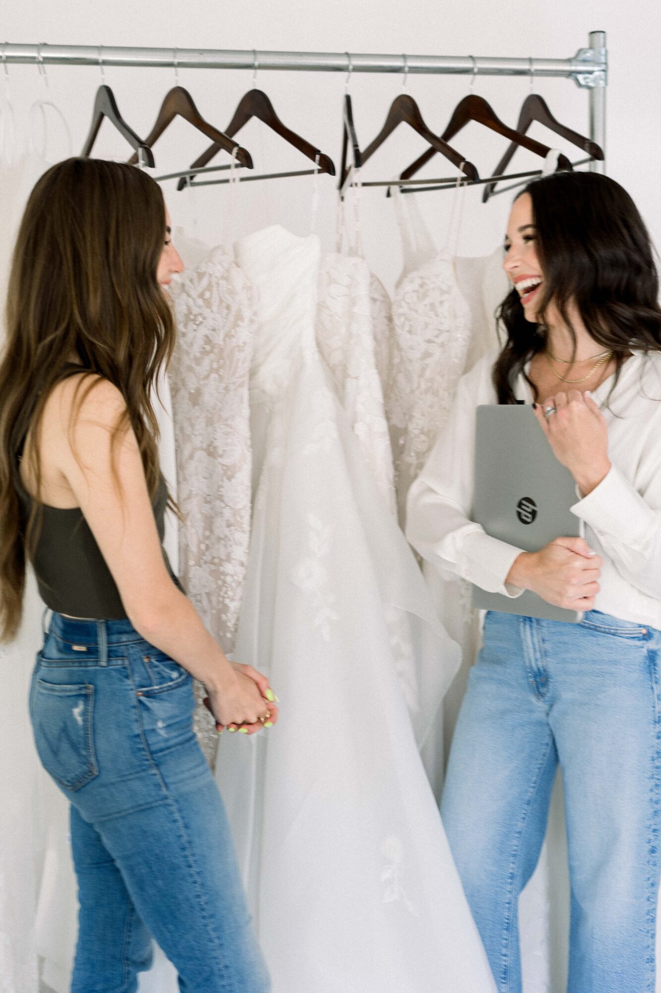 Two women smiling and talking in front of a wedding dress on a rack, holding a tablet, in a bridal shop.