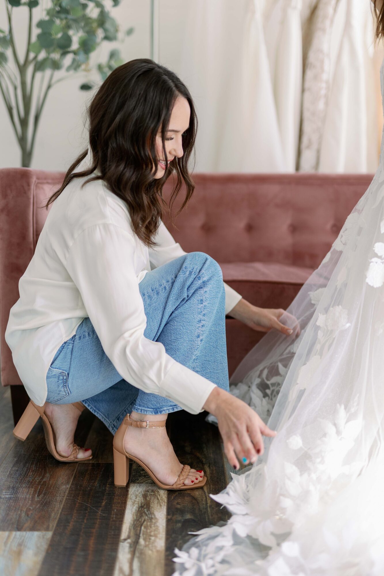 Woman crouching on floor, adjusting her shoe near a white decorative item, with a pink sofa and curtains in background.