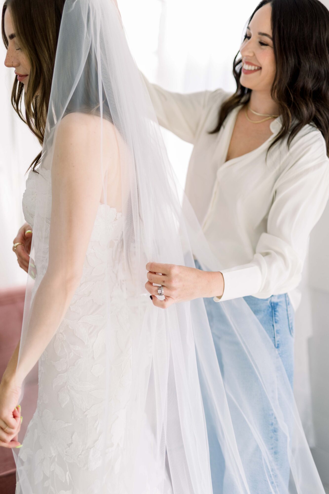 Two women, one in a wedding dress and veil, the other adjusting the veil, smiling warmly, indoors with bright lighting.
