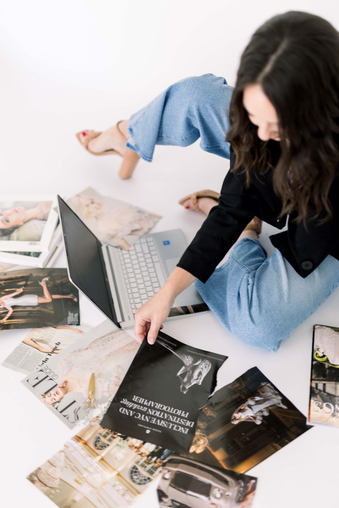 Person sitting on floor with magazines and a laptop, reaching for a magazine, with long dark hair and casual clothing.