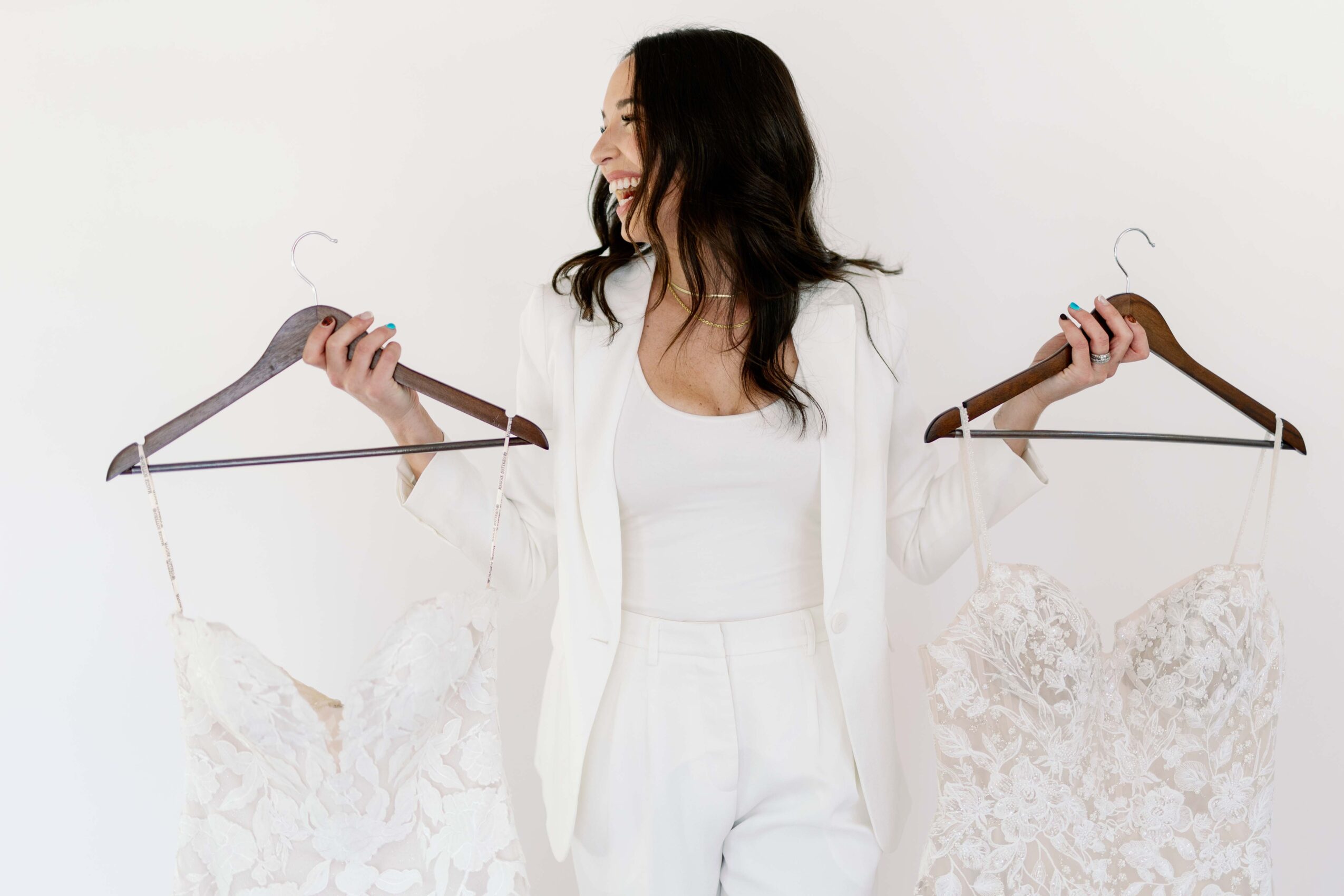 Woman with long dark hair holding two white lace dresses on hangers, standing against a white background.