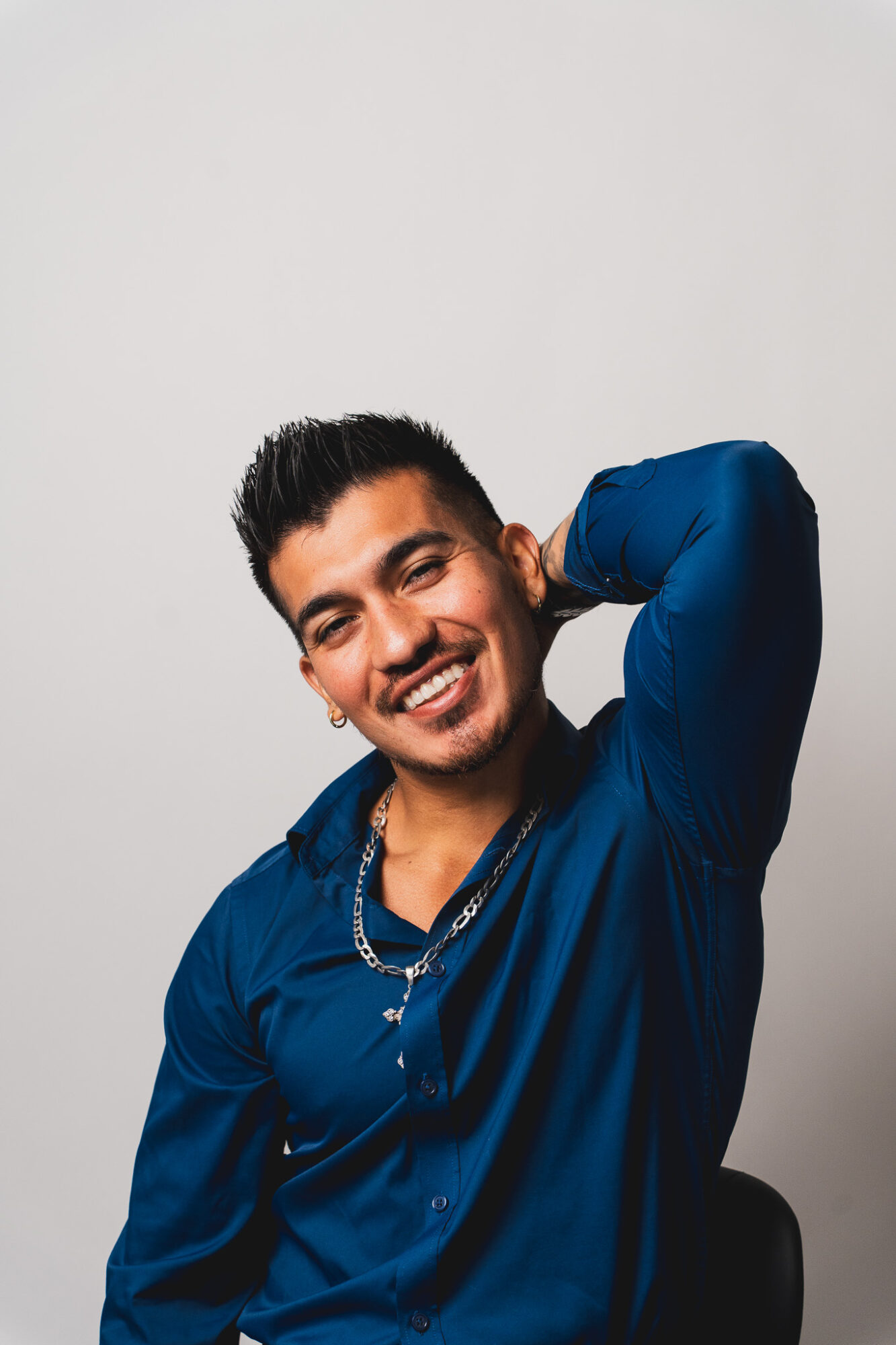 Young man smiling with one hand behind his head, wearing a blue shirt, against a plain background.
