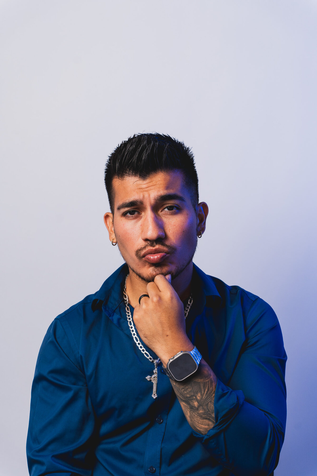 Young man with short dark hair, wearing a blue shirt, posing with hand on chin against plain background.